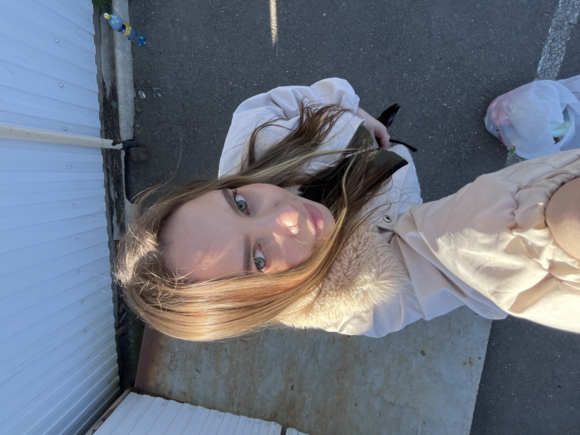 A selfie of a young woman with long blonde hair is taken from a high angle. She is wearing a light-colored jacket with a fur collar. The background includes a white metal siding wall, a grey concrete surface, and asphalt. A plastic bag sits on the asphalt. A bottle and a tool are visible near the metal siding. The location is Novopokrovskaya, Russia.