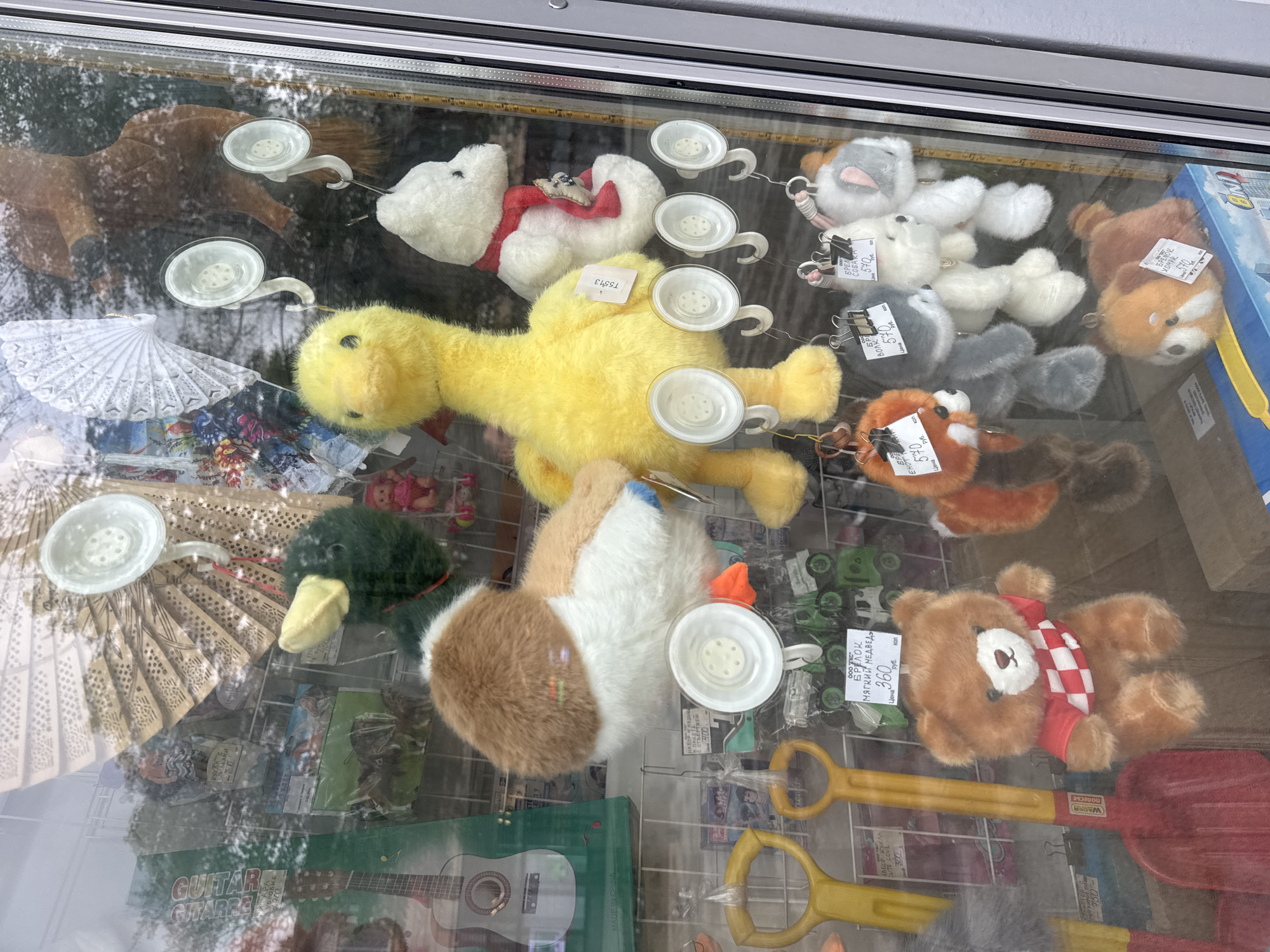 A store window display in Moscow, Russia, features a variety of plush toys. In the upper section of the display, a large yellow duck plush is prominently placed, alongside a white teddy bear with a red scarf. Behind them, a brown horse plush is visible. A plush bird with brown and white feathers is also visible. The middle section of the display shows smaller plush toys hanging from suction cups. These include a red fox, a gray wolf, a white dog, and a white cat with gray ears. On the lower shelf, a brown teddy bear wearing a red and white checkered shirt sits in the center, with yellow toy shovels to its left. A smaller plush toy resembling a red panda is on the right. The reflection on the glass shows trees and sky, suggesting an outdoor setting. The overall impression is a toy store window showcasing a colorful collection of stuffed animals.