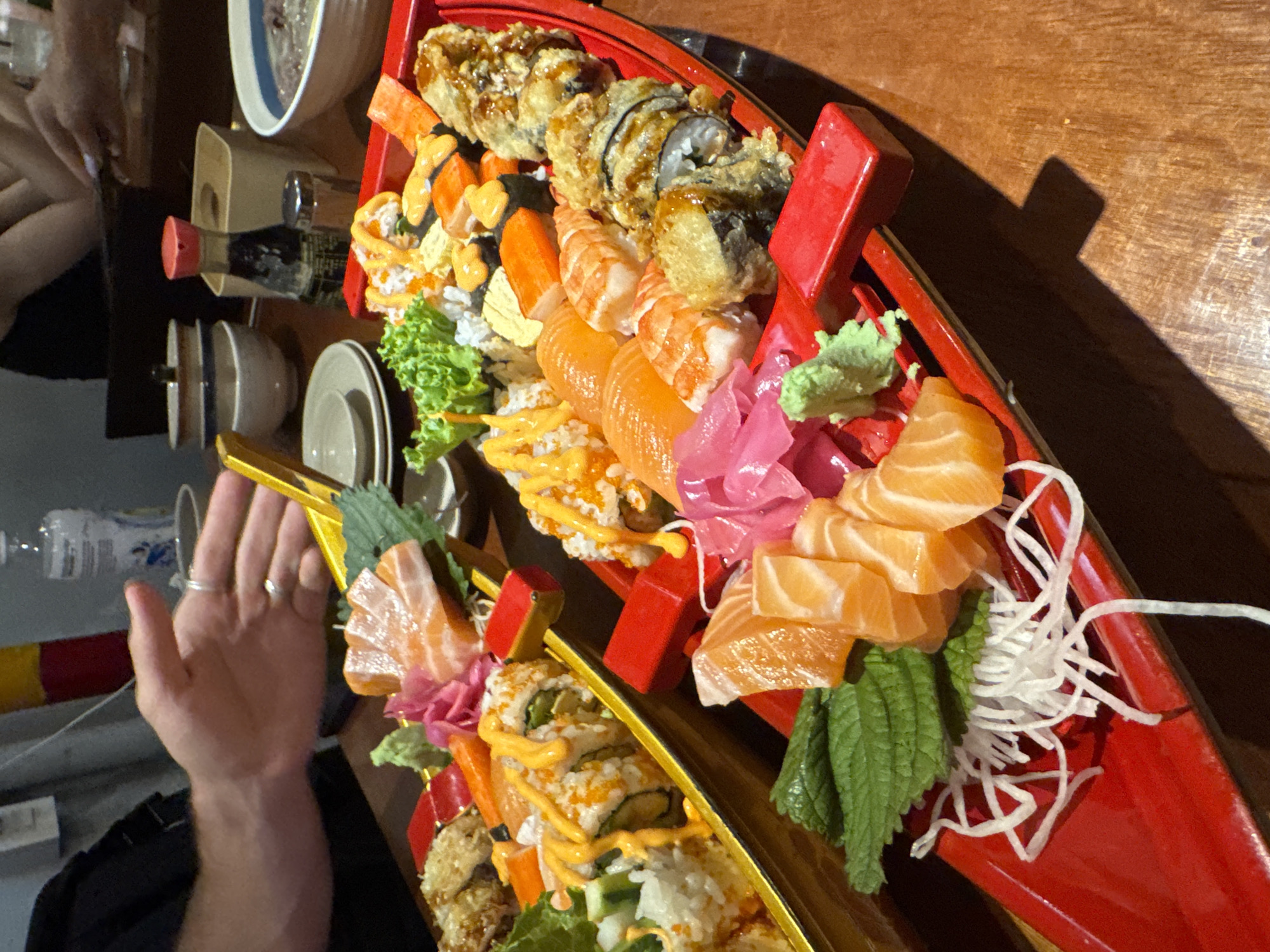 A boat-shaped platter laden with sushi and sashimi sits on a wooden table. The platter is divided into red and gold sections, each holding a variety of sushi rolls, including tempura rolls, and salmon sashimi. Garnishes include wasabi, pickled ginger, shredded daikon radish, and green leaves. A person's arm and hand are visible near the platter. Also visible are a bowl, a small soy sauce bottle, small sauce dishes, and a bottle of water. The environment suggests a restaurant setting. The location is Đà Nẵng, Vietnam.