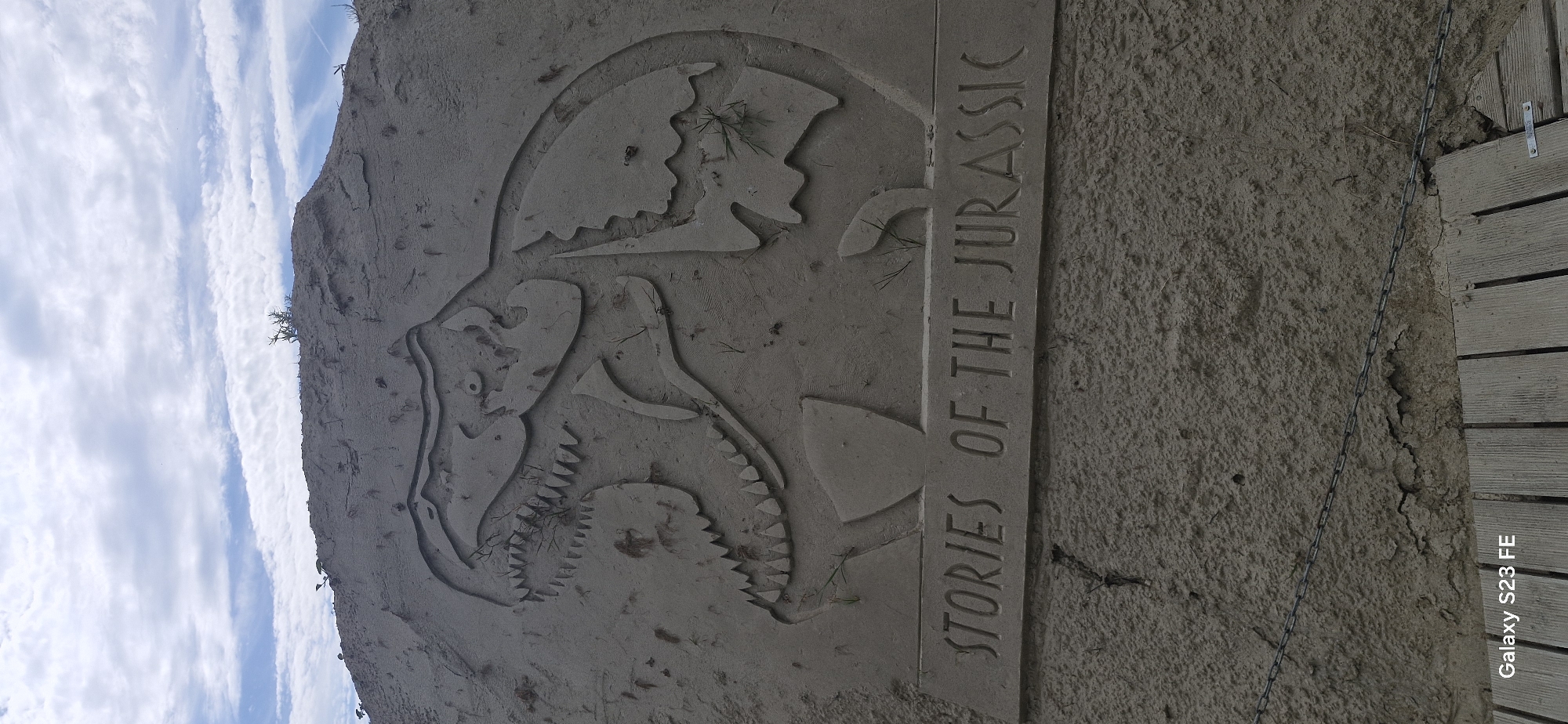 The image shows a sand sculpture of a Tyrannosaurus Rex with the words "STORIES OF THE JURASSIC" carved into a plaque below it. The sculpture is located outdoors, on a sandy embankment. The sky is visible in the background with some clouds. There are some sparse weeds and dry grass growing on and around the sculpture. A thin metal chain runs horizontally across the lower part of the sand sculpture, and below that are some wooden planks. The bottom left corner of the image has a watermark that reads "Galaxy S23 FE". The overall scene appears to be a park or an outdoor exhibition space.