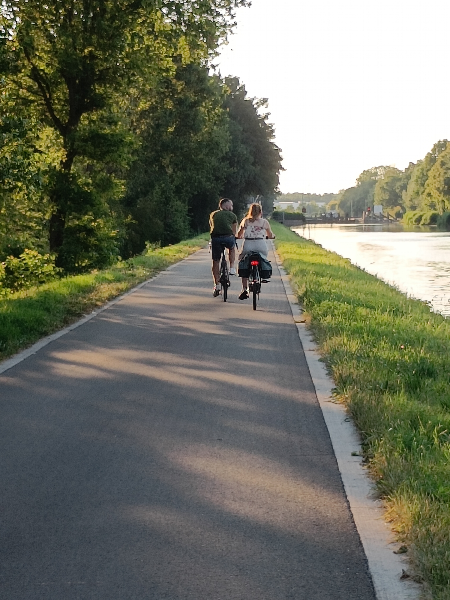 In the city of Haacht, Belgium, a paved pathway runs alongside a tranquil canal. The path is lined with lush green grass and mature trees casting dappled shadows. Two individuals are cycling away from the viewer on the pathway. The person in front, on the left, is a male wearing a green t-shirt and dark shorts. He is on a black bicycle. The person behind him, on the right, is a female wearing a white patterned top and light-colored shorts. She is also on a black bicycle, which has panniers attached to the rear rack. The canal reflects the bright sky, and distant structures, possibly a bridge or lock system, are visible across the water. The scene suggests a peaceful recreational activity in a natural setting.