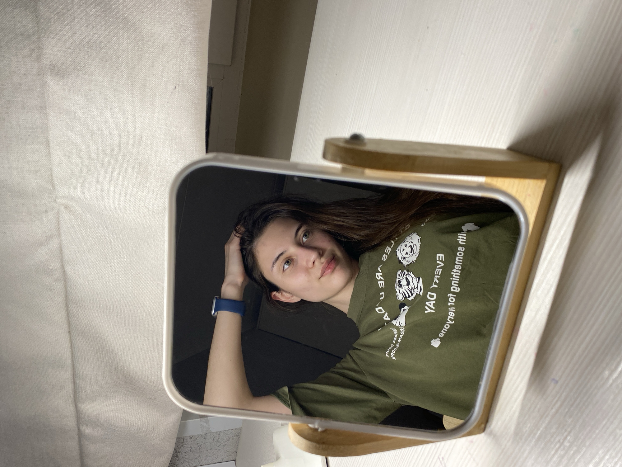 A young woman is reflected in a small, square mirror with a light-colored wooden stand. The mirror sits on a white surface, likely a desk or wall. She is wearing a dark green t-shirt with white graphics and text that is mirrored. A blue smartwatch is visible on her left wrist. Her right hand is resting on her head. The background behind the mirror shows a section of a light-colored wall with a curtain to the left.
There is no evidence in the photo to determine the location is Votkinsk, Russia.