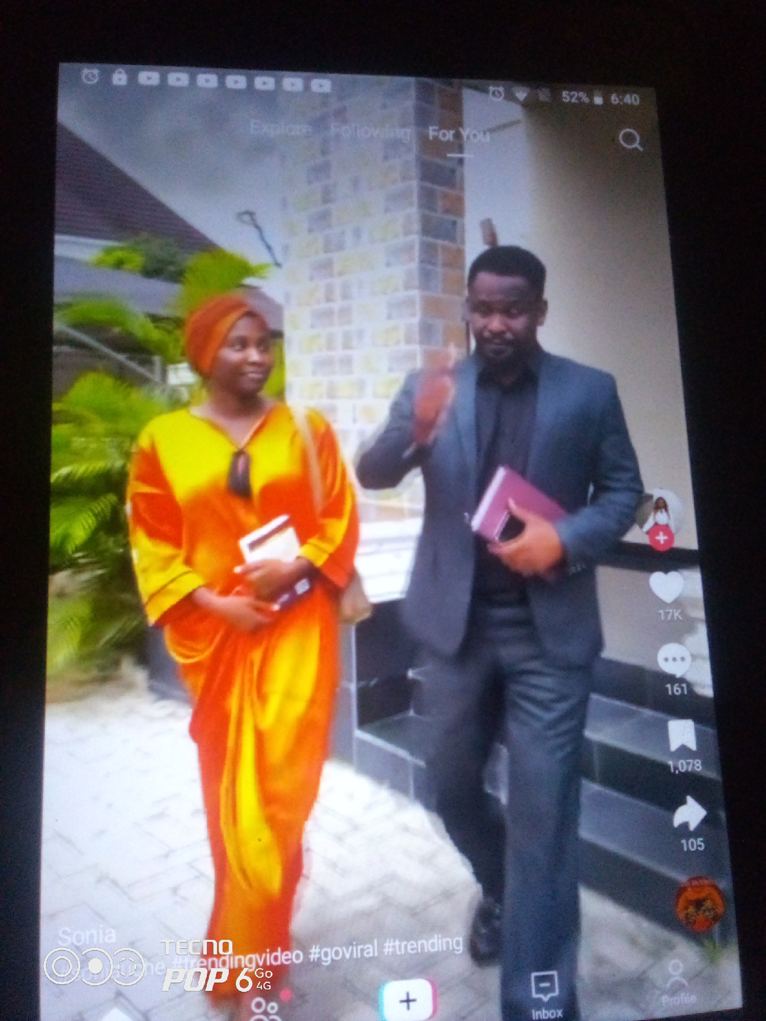 The image shows a screenshot of a video from a mobile device, possibly a TikTok interface. Two individuals are depicted walking together. On the left, a woman wears an orange and yellow patterned outfit and a headscarf. She is holding a book. On the right, a man in a dark suit and shirt is also holding a book. The background appears to be an outdoor setting with a brick wall and greenery. Overlayed text includes "Explore Following For You", "52% 6:40", and social media interaction icons with numbers like "17K", "161", "1,078", and "105". At the bottom of the screen, text reads "Sonia TECNO POP 6 4G #trendingvideo #goviral #trending" with icons for a plus sign, "Inbox", and "Profile". The device is displaying information consistent with a smartphone. The location of Howayi, Nigeria is not ascertainable from the image content itself.