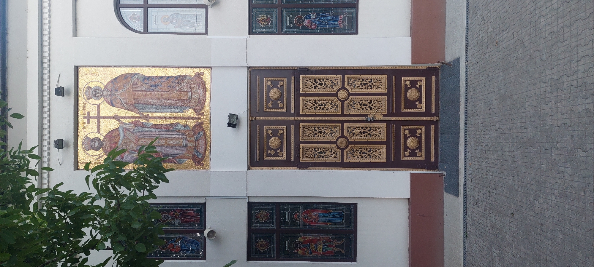 The image shows the facade of a building, likely a church, located in Iași, Romania. The central feature is a large, ornate, dark brown double door with intricate gold metallic detailing. Above the door, a large mosaic depicts two figures in traditional religious attire holding a cross. To the left and right of the mosaic and door, there are stained-glass windows featuring religious imagery. A tree with green leaves partially obscures the upper left section of the building. The ground in front of the building is paved with cobblestones. The overall impression is of a well-maintained religious structure with traditional architectural and artistic elements.