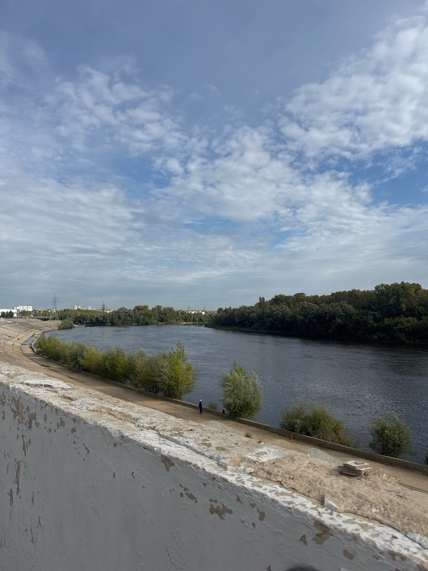 The image shows a view of the Belaya River in Ufa, Russia. The river is wide and dark, with a forested bank on the right side of the image and a concrete embankment on the left. A few trees are scattered along the embankment, and a single person in a white hat is standing near the water's edge. In the distance, the skyline of Ufa is visible, with buildings and power lines. The sky is cloudy, with patches of blue peeking through. The foreground is dominated by a rough, white concrete wall.