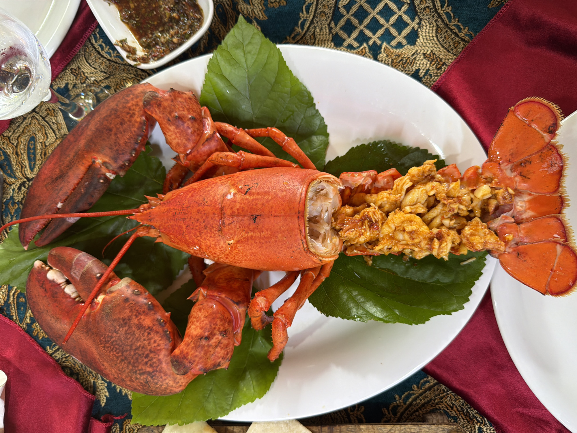 An overhead shot shows a cooked lobster presented on a white plate, adorned with large green leaves. The lobster is a vibrant orange-red color, with its claws and tail prominently displayed. The body of the lobster has been split lengthwise and filled with a mixture of what appears to be finely chopped meat and spices. In the background, a glass of clear liquid, possibly water or wine, sits on a patterned tablecloth in shades of teal and gold, with a section of a dark red napkin visible. To the right of the plate, a small white dish contains a dark, textured sauce. The lighting suggests an indoor setting, possibly a restaurant. The image does not provide any specific information about the city of Kemer, Turkey, or any individuals present.