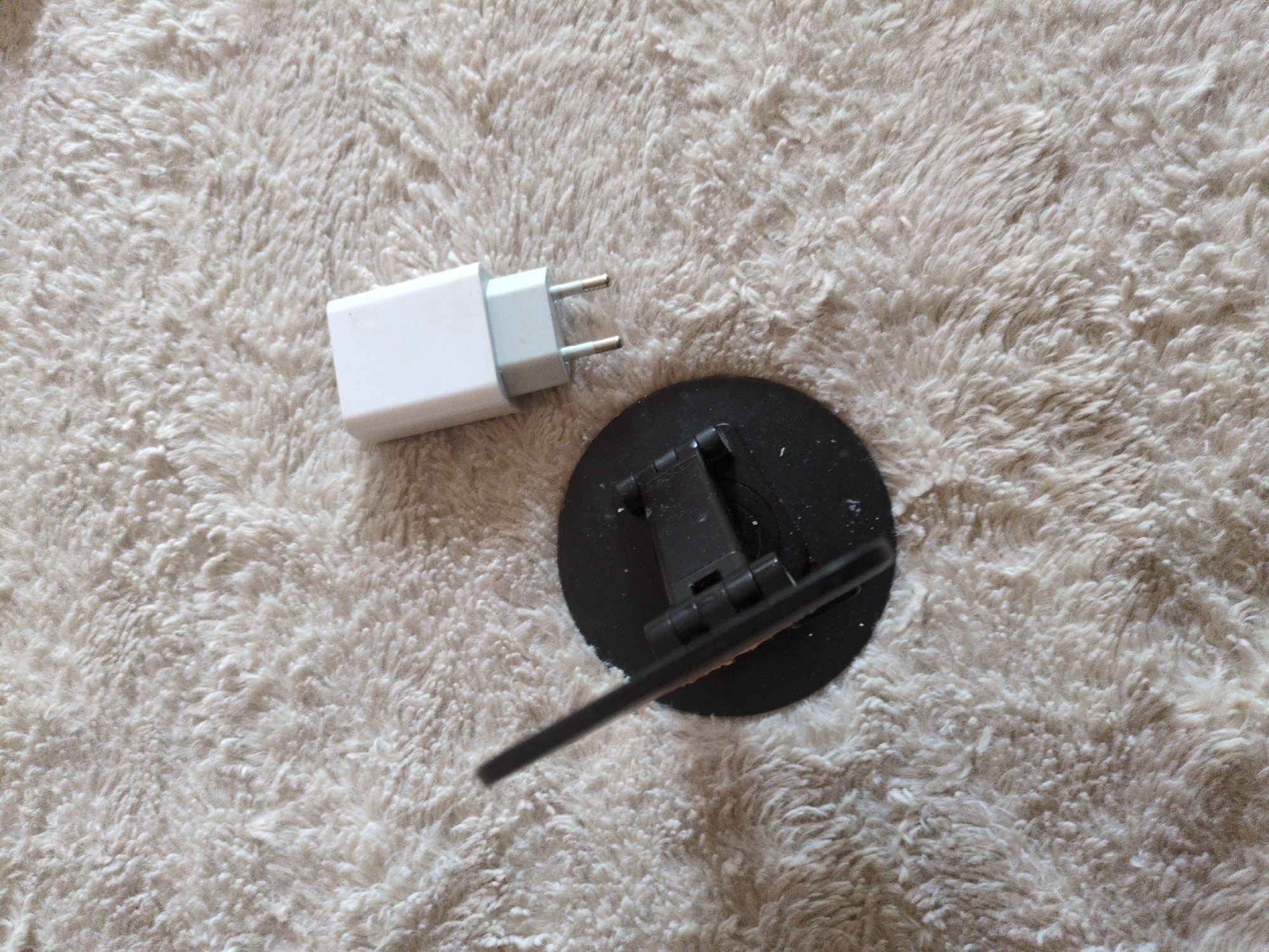 An overhead shot shows a white power adapter and a black stand for an antenna, both resting on a fuzzy, light-colored carpet. The power adapter, a common phone charger, is positioned in the upper right quadrant of the image, with its prongs pointing downwards. To the left of the adapter is the black stand, which has a circular base and a hinged component that supports a thin, black antenna. The antenna is angled upwards and to the left, its form partially obscured by the stand. The carpet's texture is a key feature of the scene, its shaggy fibers creating a soft, neutral backdrop for the electronic accessories. The lighting is even, with no harsh shadows or direct glare, suggesting an indoor environment without prominent light sources.