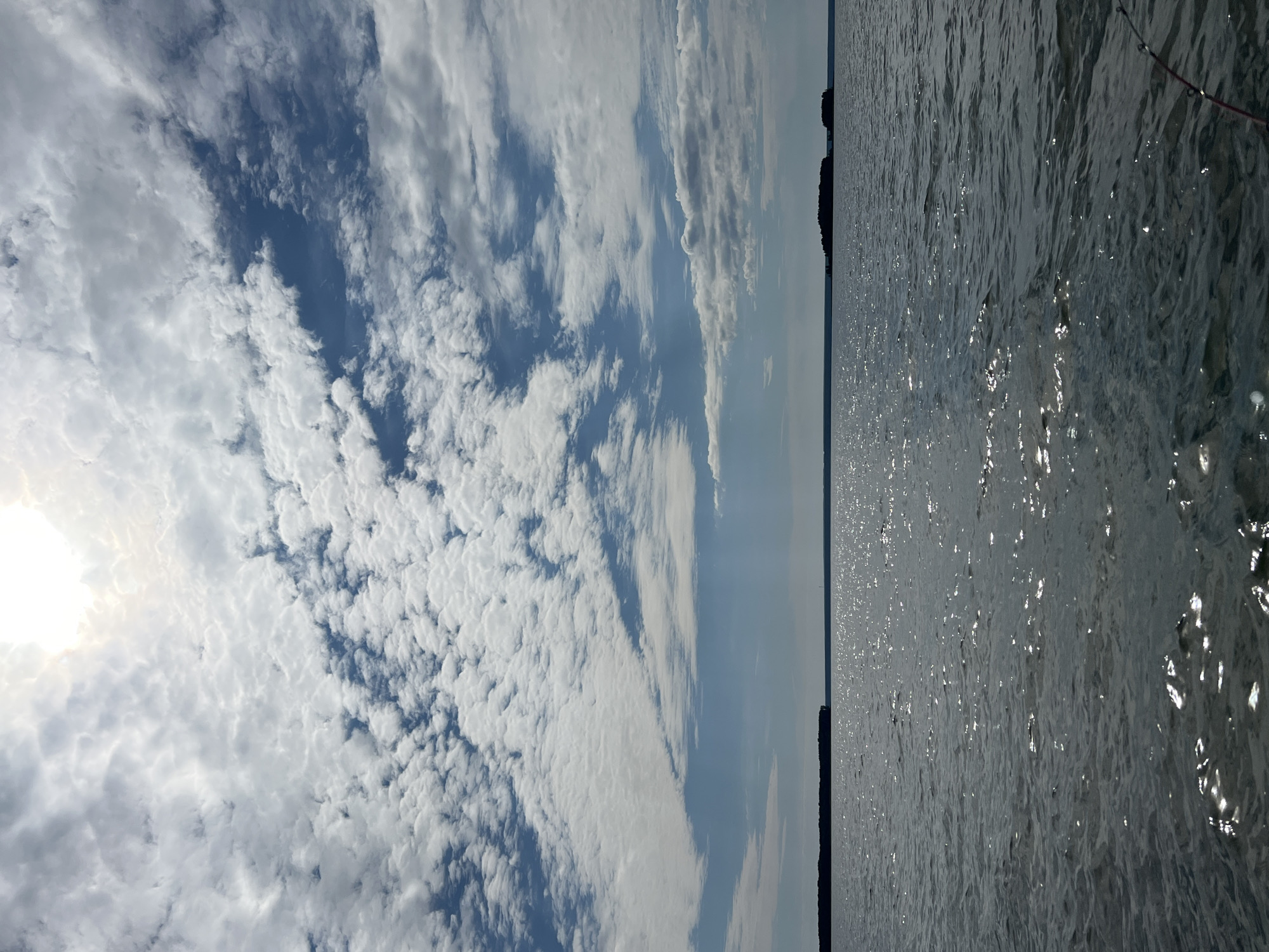 The image depicts a body of water, possibly a lake or sea, under a partly cloudy sky. The sun is visible through a break in the clouds, casting bright reflections on the water's surface. In the distance, landmasses with trees are visible along the horizon, suggesting the presence of islands or a distant shoreline. The water is calm with gentle ripples. The scene is natural and serene. The location is not specified in Novopichugovo, Russia.