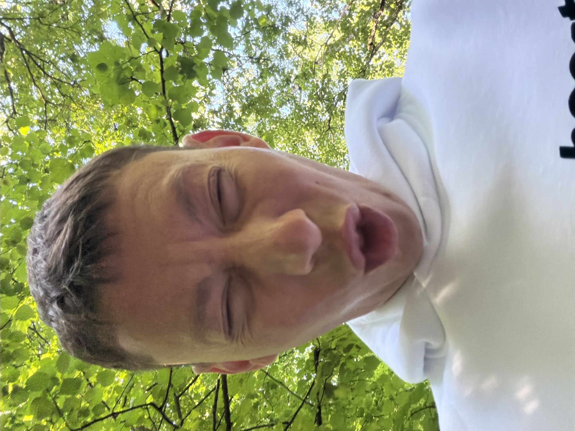 In a low-angle, close-up shot, a person with short, dark hair and a white hoodie is captured outdoors. Their eyes are closed, and their mouth is open as if in mid-sentence or expression. The background is dominated by lush green leaves of trees, with some branches visible against a bright sky. The scene appears to be set in a park or wooded area. There is no visible indication of the city of Moscow, Russia.