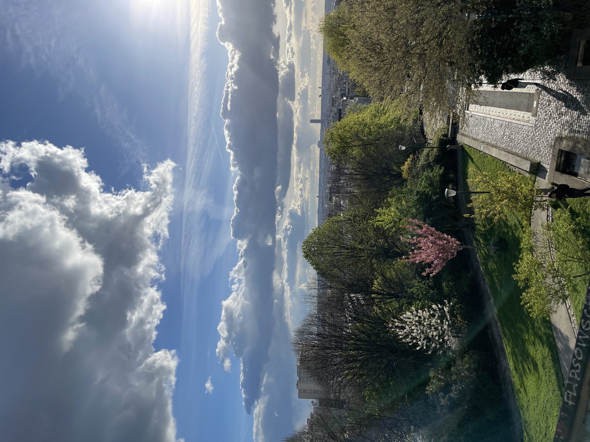 The image depicts a panoramic view of Paris, France, likely captured from an elevated position within the Montmartre district, such as Square Louise-Michel or Square Marcel Bleustein-Blanchet. The scene unfolds under a dynamic sky featuring large, bright cumulus clouds contrasted with darker, shadowed cloud masses and patches of clear blue. Strong sunlight originates from the upper right, casting long shadows.
In the immediate foreground, at the bottom left, a dark wall displays white graffiti reading "FLIPSOWSCH". A multi-tiered pathway, constructed of grey cobblestones with a central section of parallel metal grates, descends through a terraced garden. One individual, dressed in dark clothing and light pants, walks down this cobbled path in the lower right, casting a long, distinct shadow. Another individual, also in dark attire, is partially visible at the bottom left, near the graffiti wall. Several dark lampposts are positioned along the path.
The midground is dominated by a verdant park or garden with lush green grass and numerous trees. These trees exhibit varying stages of spring foliage: some are bare-branched, while others display vibrant new green leaves. Distinctly, one tree with white blossoms and another with pink blossoms are visible.
Beyond the park, the urban sprawl of Paris extends to the horizon. The cityscape consists primarily of dense, low-to-medium-rise buildings, characteristic of Parisian architecture. The Montparnasse Tower, a tall, dark skyscraper, stands prominently in the distant center-right of the skyline. Other distant structures are visible through a slight atmospheric haze.