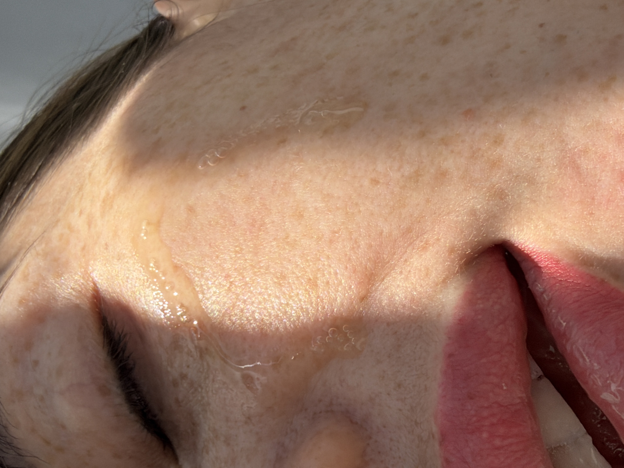 The image displays a close-up, vertical shot of the right side of an individual's face, observed from approximately the forehead/temple area down to the mouth. The subject has light-toned skin with numerous scattered freckles, particularly prominent on the cheek and around the eye. The right eye is mostly closed, with dark eyelashes visible. Clear, viscous liquid is present on the skin below the eye, forming distinct droplets and streaks, suggesting a flow down the cheek. The right corner of the mouth is visible, slightly open, revealing a portion of the reddish-pink inner lip and a glimpse of the tongue. Dark brown hair is visible along the upper right edge of the frame, near the temple. Strong directional lighting illuminates the face, creating a shadow across the upper left portion of the eye and nose area. The background is uniformly light grey and out of focus, providing no contextual information. There are no discernible elements within the image to indicate a specific location, including the city of Saint Petersburg, Russia.