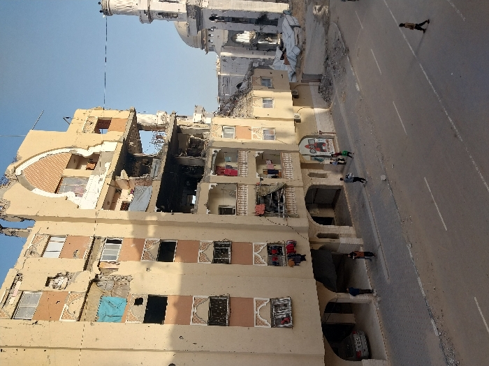 The image depicts a street scene in Khan Yunis, Palestinian Territory, characterized by significant urban destruction. In the foreground, a multi-story, light-colored residential or mixed-use building dominates the left, exhibiting extensive structural damage. Multiple floors show large sections of exterior walls missing, exposing interiors, with visible rebar and numerous pockmarks across its facade. A large blackened void indicates severe impact and fire damage on the middle-upper floors. Blue and white tarps cover openings on different levels, while clothing hangs on several damaged balconies. The ground floor features arched openings, one housing a silver sedan, with three adults present on the adjacent paved sidewalk.
To the right of the damaged building, a mosque with a distinctive minaret and dome is visible. The minaret is partially destroyed, lacking its upper spire section, and surrounded by rubble. Additional collapsed structures and debris are visible between the main building and the mosque.
The foreground shows a paved street with white lane markings, partially covered in sand and small debris. A paved sidewalk runs parallel to the building. On the sidewalk, near the center, three children are visible, two in green shirts. Further right, an adult male walks on the street, away from the viewer. The sky is clear blue.