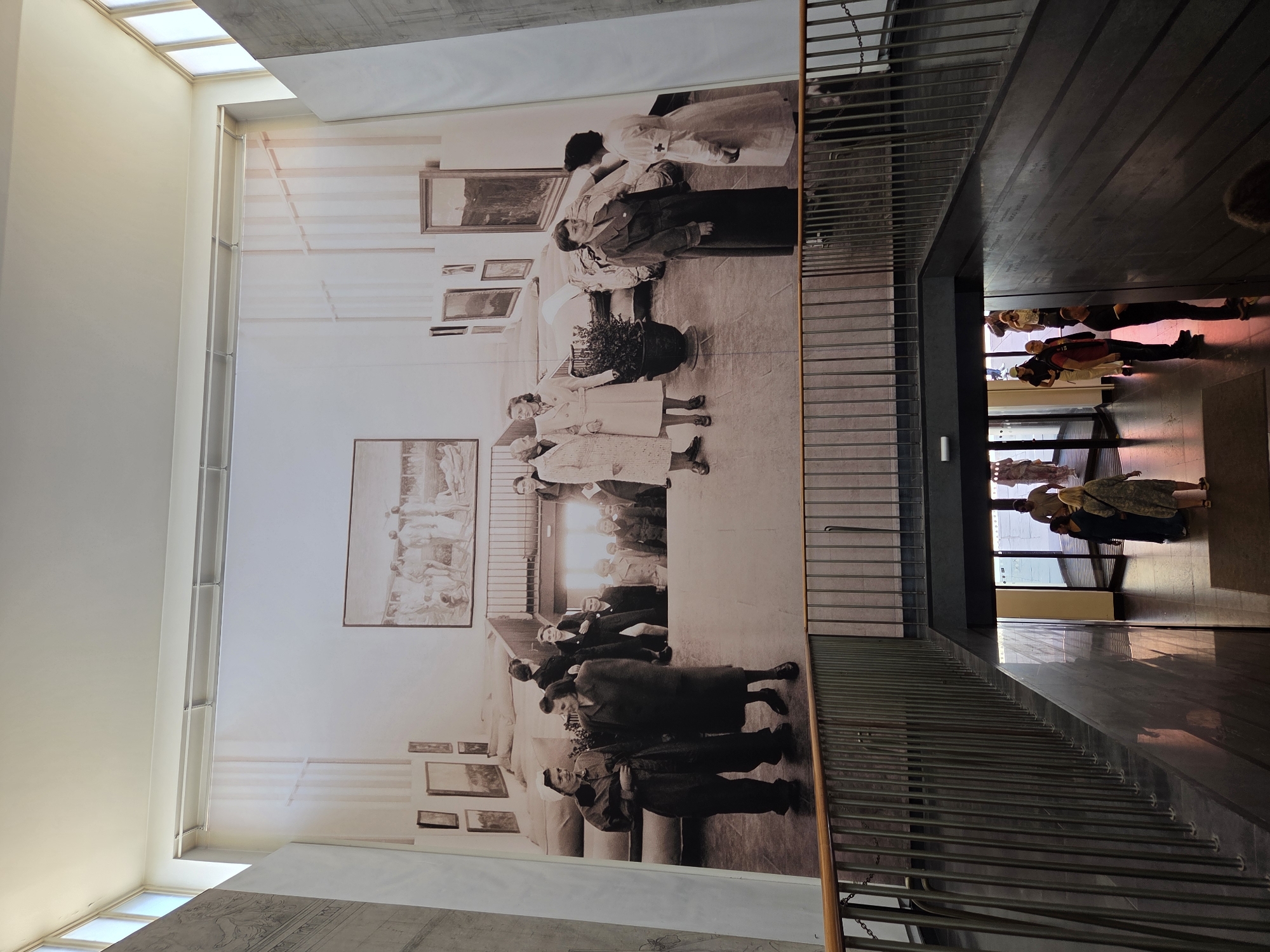 The image shows a large mural on a wall depicting a historical scene with several people. The mural is in black and white and appears to be a photograph. In the foreground, there is a staircase with a metal railing leading down. Below the mural, in the background, real people are walking through a doorway and down a hallway. The environment appears to be an indoor public space, possibly a museum or gallery.
The building is Moderna Museet Malmö in Malmö, Sweden. The mural is a large-scale artwork that reproduces a photograph of the opening of the Moderna Museet in Stockholm in 1958. It shows visitors, including artists and cultural figures, attending the inauguration. The Moderna Museet Malmö is located in a former power station, and the architectural style of the building is evident with its concrete walls and large windows.