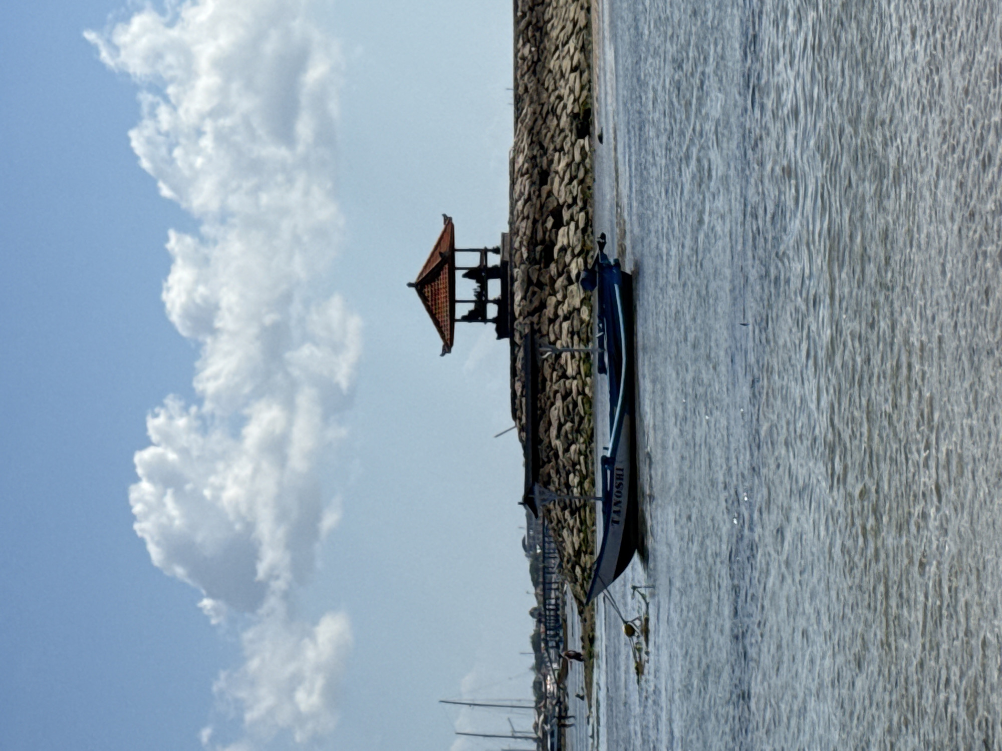 The image depicts a tranquil scene at a coastal location in Denpasar, Indonesia. In the foreground, the gentle waves of the ocean create a shimmering effect on the water's surface, with the seabed visible beneath in shallow areas. A blue and white boat named "TANOSHI" floats on the water, tethered by a rope. In the mid-ground, a man-made stone breakwater extends into the sea, topped by a traditional Balinese pavilion with a red tiled roof. The pavilion appears to have people sheltered within it. Further in the background, several sailboats are anchored, and a distant coastline with buildings is visible under a bright blue sky with scattered white clouds. The overall atmosphere is one of peaceful coexistence between man-made structures and the natural coastal environment.