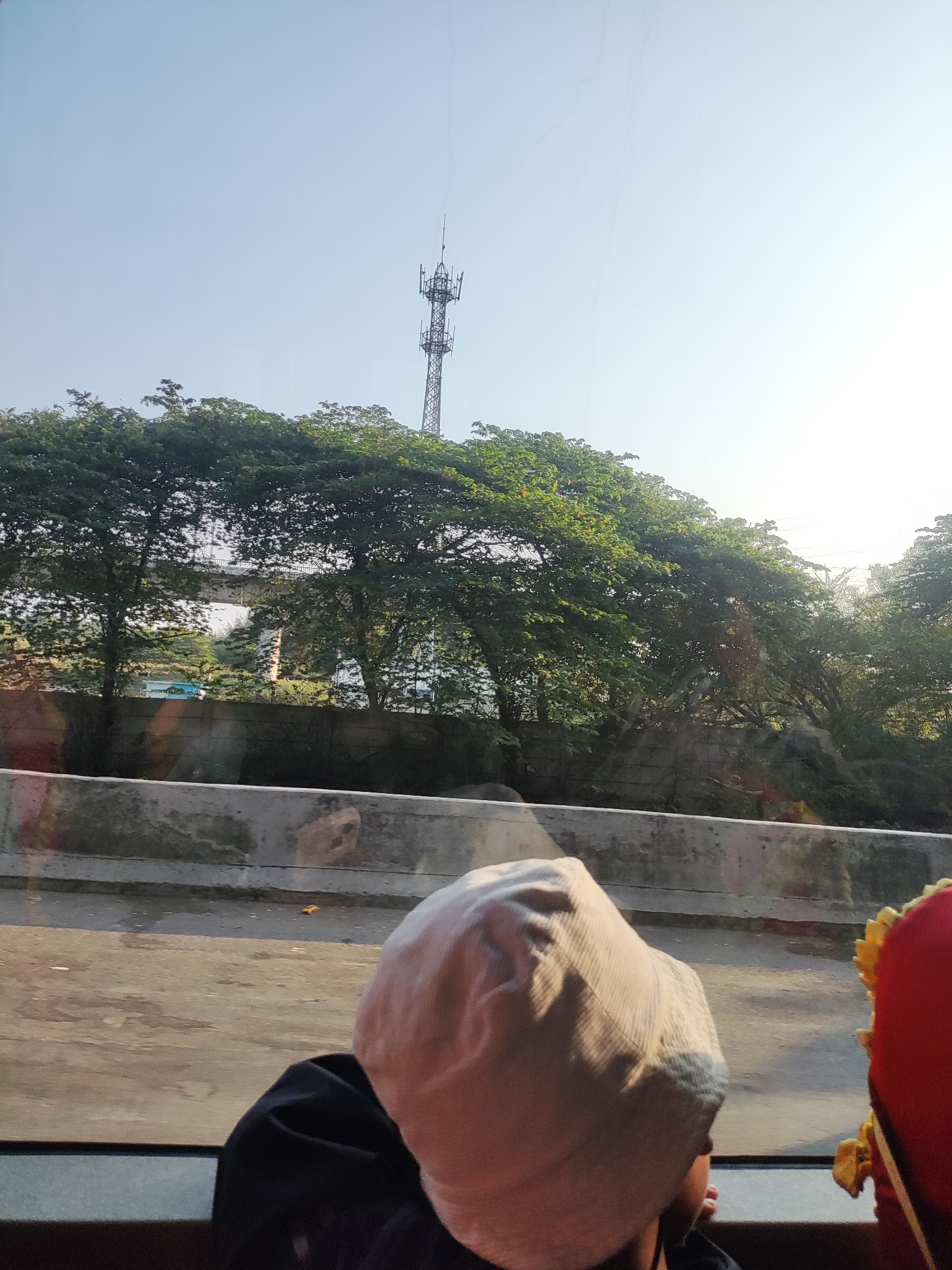 The image shows a view from inside a vehicle, looking out at a telecommunications tower in the distance. The tower is tall and skeletal, with multiple antennas at its top. In the foreground, the back of a person's head is visible, wearing a light-colored hat. To the right, another person's head is partially visible, wearing a red hat with yellow trim. The scene appears to be outdoors, with lush green trees lining the horizon and a concrete barrier in the middle ground. The sky is clear and bright. The specific location in Hegarmukti, Indonesia, is not discernible from the image itself, but the presence of a telecommunications tower and the overall vegetation suggest an urban or semi-urban environment.