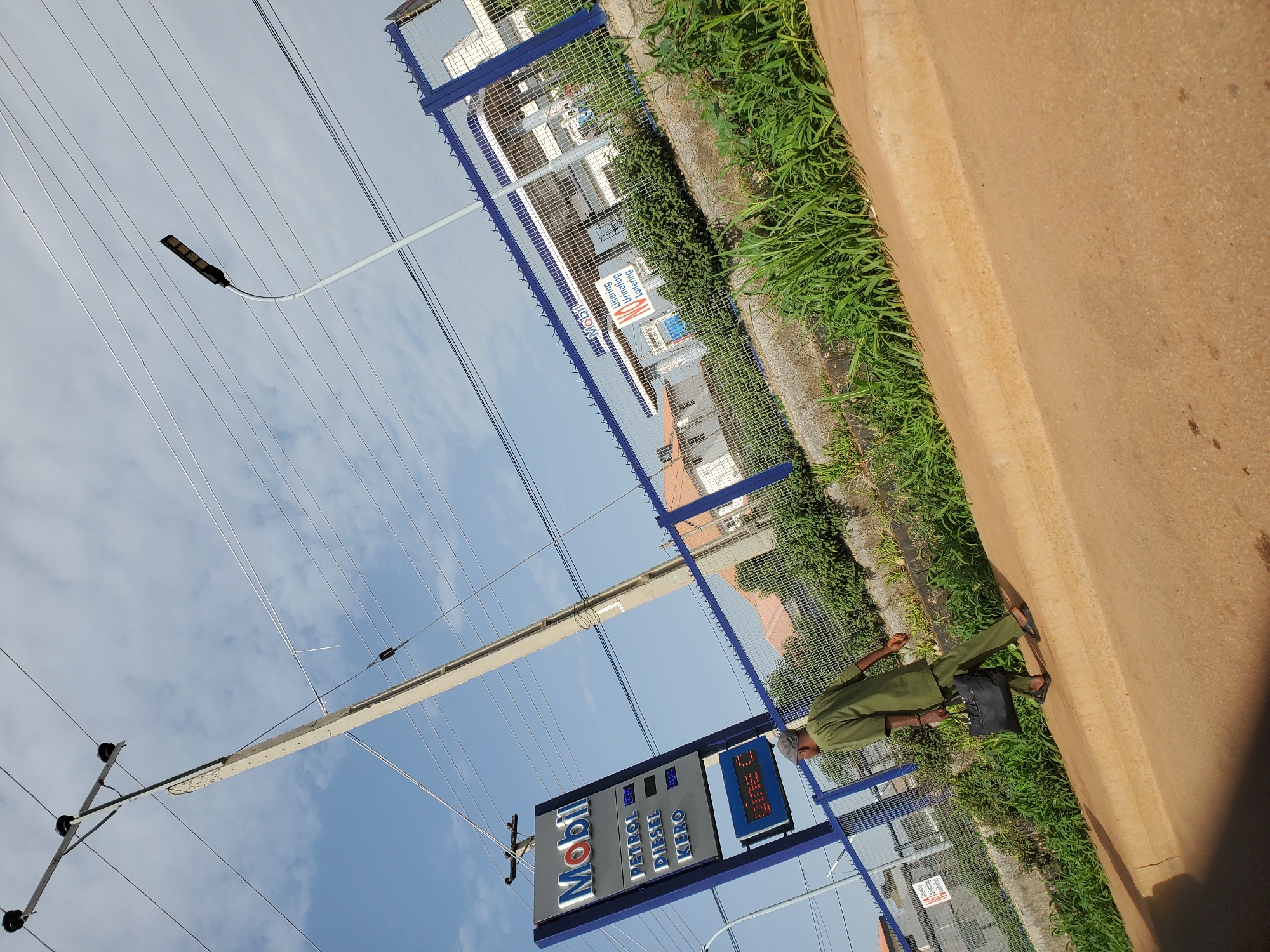 The image captures a street scene in Bukuru, Nigeria, dominated by a tall concrete utility pole with multiple wires stretching across the frame against a cloudy blue sky. To the left, a tall, blue and silver Mobil petrol station sign displays prices for petrol, diesel, and kero, with a red illuminated section at the bottom. A man dressed in a green traditional outfit, carrying a black briefcase, walks away from the sign on a dirt path beside a curb. In the background, a blue metal fence encloses a Mobil petrol station, with buildings and more greenery visible beyond. A modern street lamp is mounted on a curved pole, also visible against the sky. The overall scene depicts a typical urban environment with commercial signage, infrastructure, and pedestrian activity.