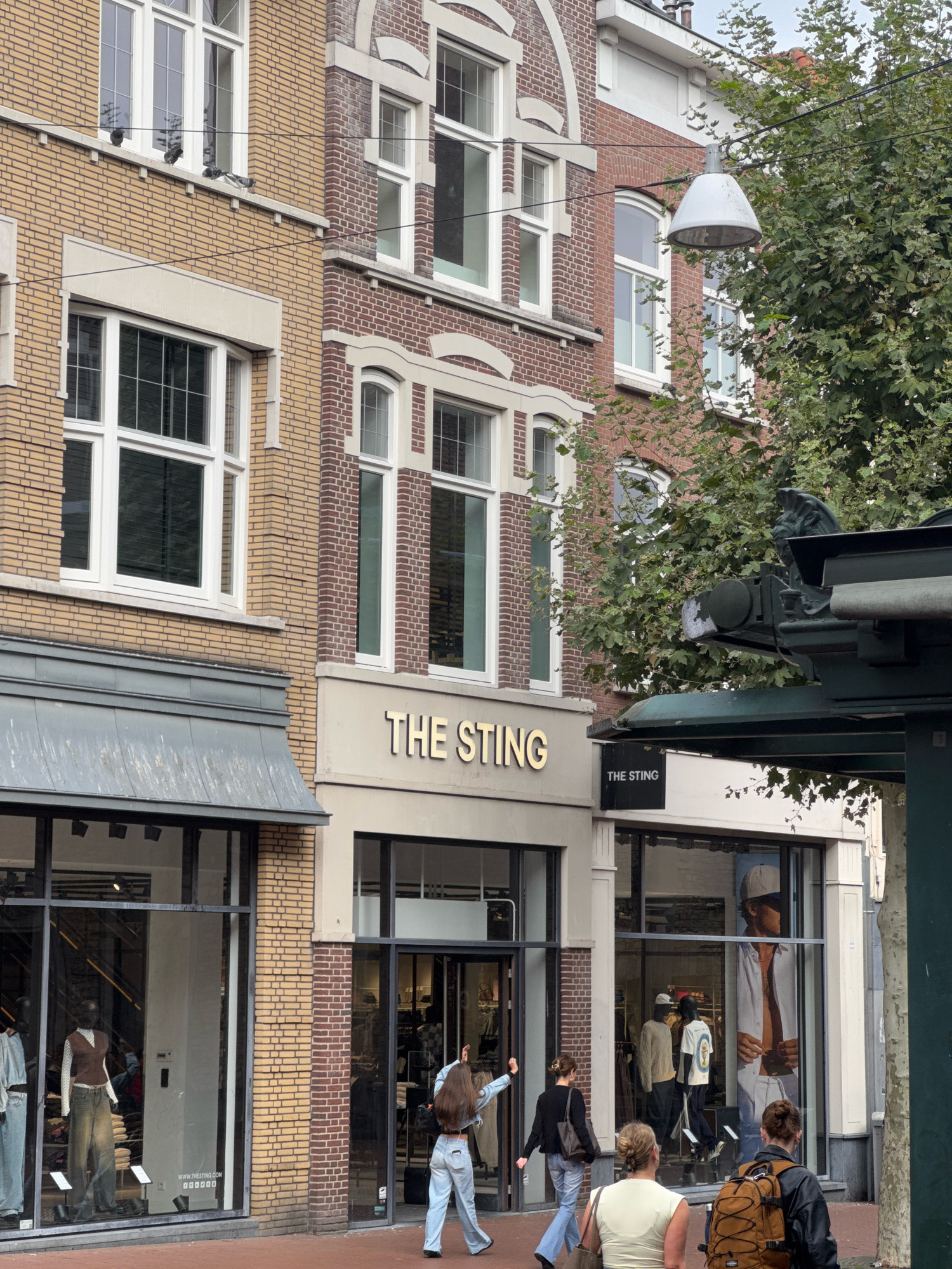 The image shows a street scene in Nijmegen, Netherlands, with two storefronts belonging to "The Sting" clothing store. The building facade is brick and stone. In the foreground, several people are walking on the sidewalk. On the left, a person is walking away from the camera, looking into the window display of the first "The Sting" store. The window displays mannequins wearing casual clothing. To the right, two women and a man are walking away from the camera. The man is wearing a backpack. The second "The Sting" store has a window display featuring a large poster of a man. A street lamp hangs from wires overhead. Trees with green leaves are visible in the background.