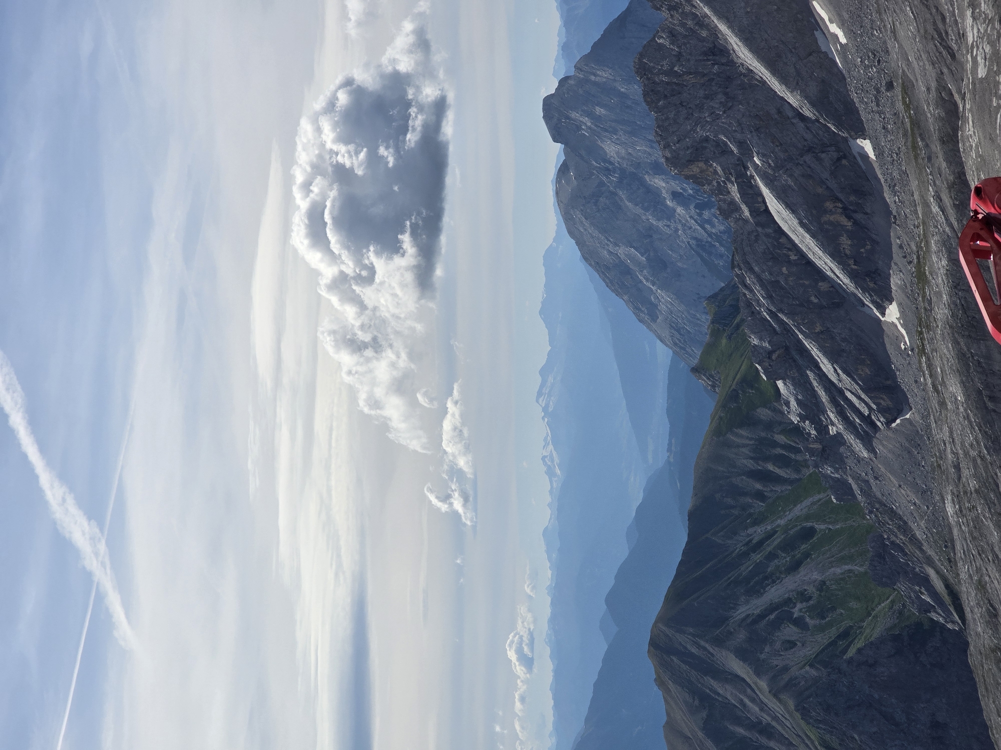 The image depicts a panoramic view of the Bavarian Alps, likely from a high vantage point on the Zugspitze, Germany's highest peak, which is near Garmisch-Partenkirchen. The foreground is dominated by rocky, grey terrain, characteristic of alpine environments. A bright red object, partially visible and appearing to be a piece of equipment or machinery, is situated in the lower right corner of the frame.
The mid-ground features several layers of rugged mountain ranges. The closest mountains are jagged and steep, with patches of green vegetation and scattered snow on their slopes. Further into the distance, the mountains become softer in contour and appear in shades of blue due to atmospheric perspective, suggesting a considerable depth of field. Snow-capped peaks are visible on the horizon, indicating the high altitude of the region.
The sky is a mix of blue and grey clouds. A prominent, large cumulus cloud formation is centrally located in the upper half of the image. Two contrails, remnants of aircraft, are visible in the upper left quadrant of the sky, forming an X shape. The lighting suggests it is daytime, with the sun likely diffused by the clouds, creating a somewhat muted light across the landscape. No people are clearly visible in the scene.