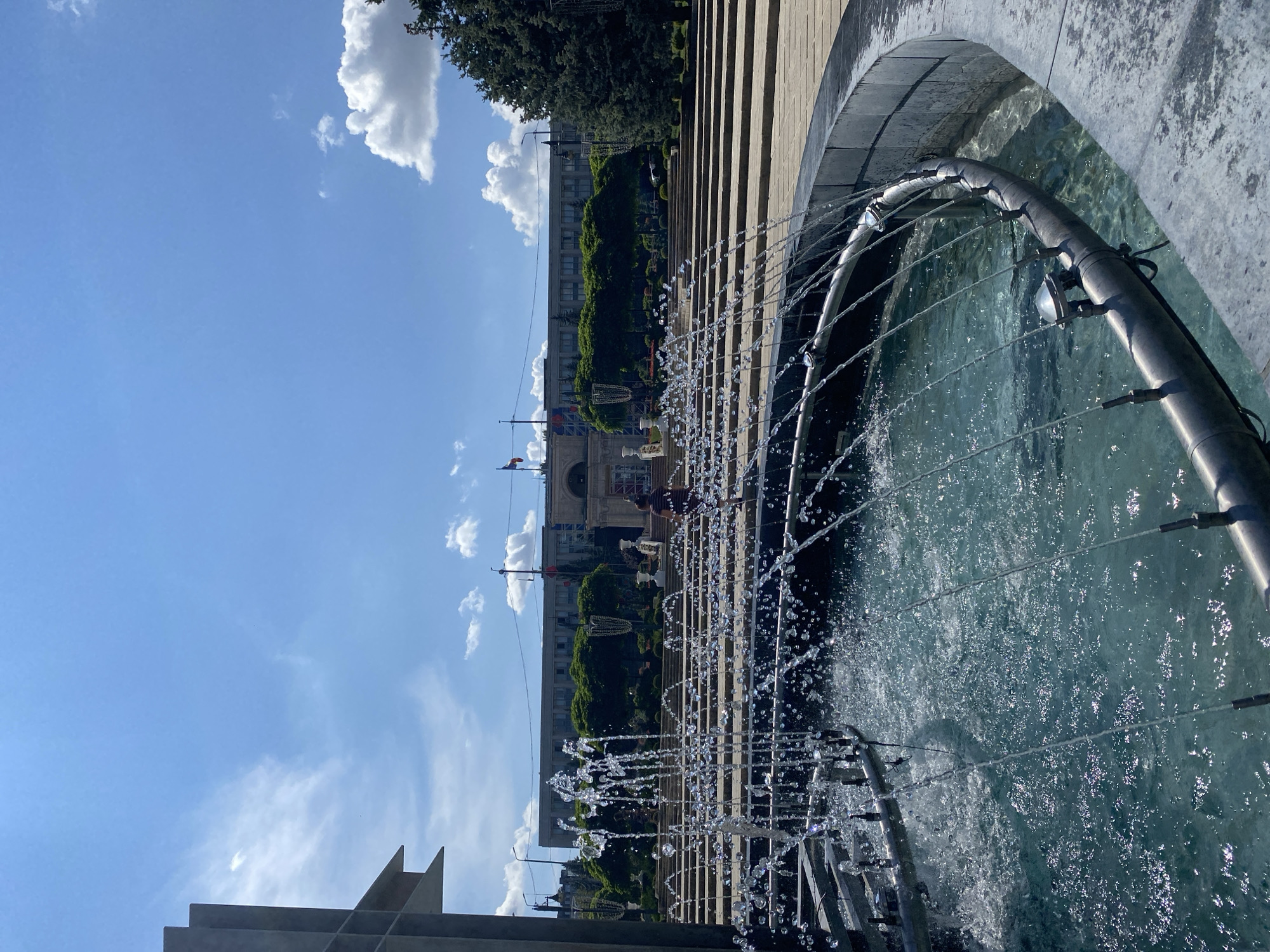 The image shows a large, multi-tiered fountain in the foreground with water spraying upwards. In the middle ground, there are a series of wide stone steps leading up to a grand building with flags and trees. The building appears to be Government House in Chișinău, Moldova, a prominent landmark. The sky is blue with scattered white clouds, indicating a clear day. A person is visible walking up the steps in the middle distance. The scene is an outdoor public space, likely a plaza or park.