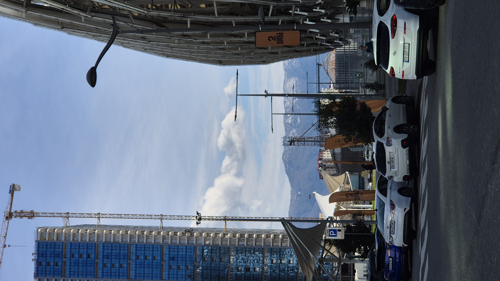 The image captures an urban scene in Batumi, Georgia, under a clear, blue sky with scattered clouds. A partially constructed skyscraper with blue glass panels dominates the left side of the frame, attended by a construction crane. In the foreground, a row of parked cars lines the right side of the street. The cars have license plates with the following alphanumeric codes: MA-012-RS, HH-629-WH, DA-880-NI, and TX-037-TT. The road stretches into the distance, leading towards a range of mountains visible on the horizon. Between the cars and the mountain backdrop, awnings and signage suggest the presence of commercial establishments. At the top of the image, sections of another building or structure overhangs the road.