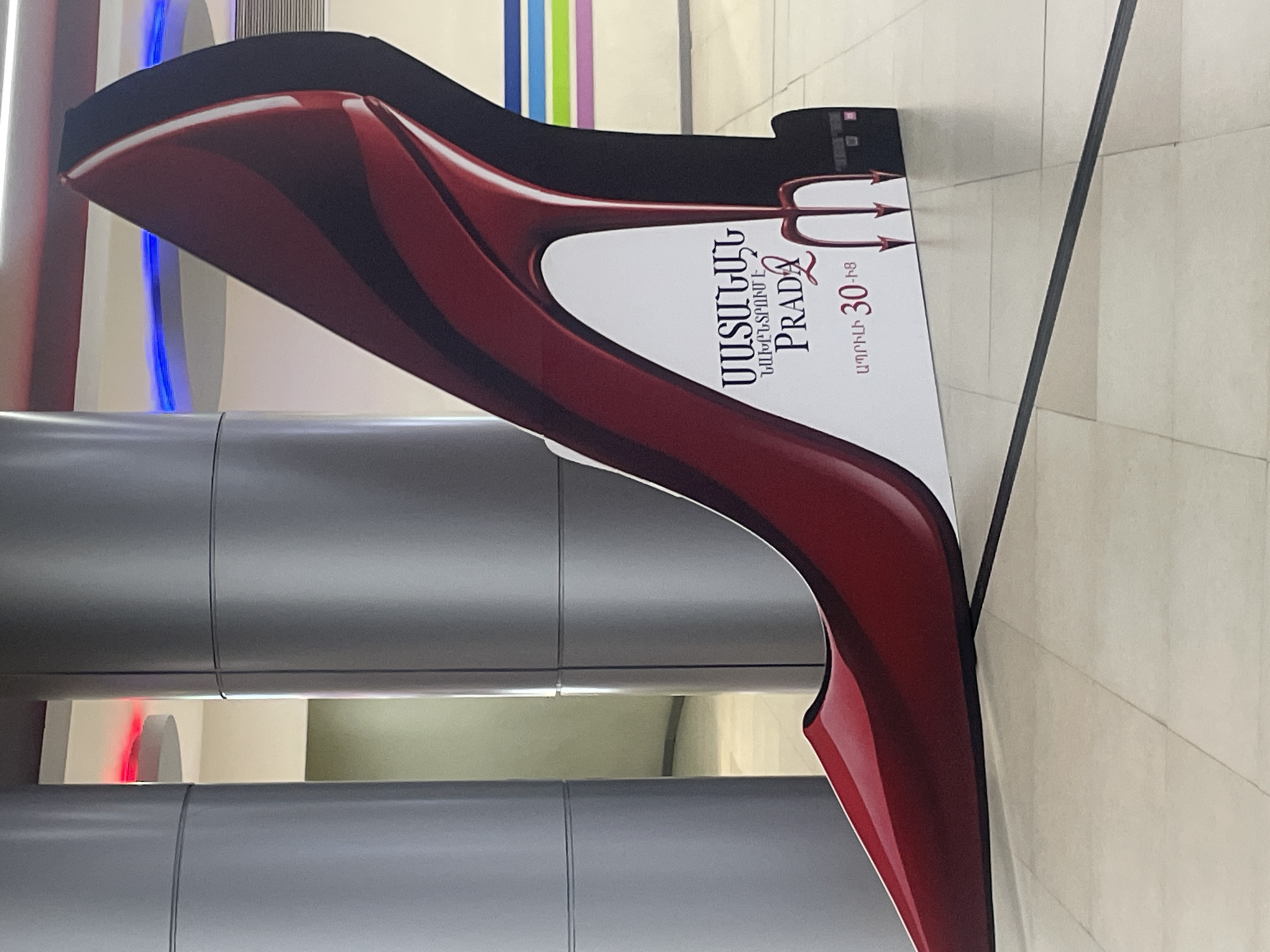 An indoor shot captures a large, vertical, stand-up advertisement shaped like a glossy, dark red and black high-heeled shoe. The design is sleek and exaggerated, featuring a pointed toe and a dramatic arch suggestive of a stiletto. A white section on the shoe's side contains text and graphics.
Prominently displayed in Armenian script are the words "ՍԱՏԱՆԱՅԻՆ ՀԱԳՆՈՒՄ Է" which translates to "Satan Wears". Below this, the brand name "PRADA" is written in a stylized font, accompanied by a small red heart icon. To the right of the brand name, a subtle devil's trident graphic is visible, reinforcing the theme. Further Armenian text below "PRADA" reads "ԱՊՐԻԼԻ 30-ից", meaning "from April 30". This indicates a promotional display for an event or collection themed around "Satan Wears Prada", starting on April 30th.
The advertisement is positioned indoors, likely within a shopping mall or large public building in Yerevan, Armenia. The background features several light gray cylindrical pillars, some with reflections of colorful lighting, including blue, red, green, and purple streaks. The floor is tiled in a light, neutral color with dark grout lines, suggesting a clean, modern commercial environment. The overall impression is one of a striking, fashion-oriented promotion in a public space.