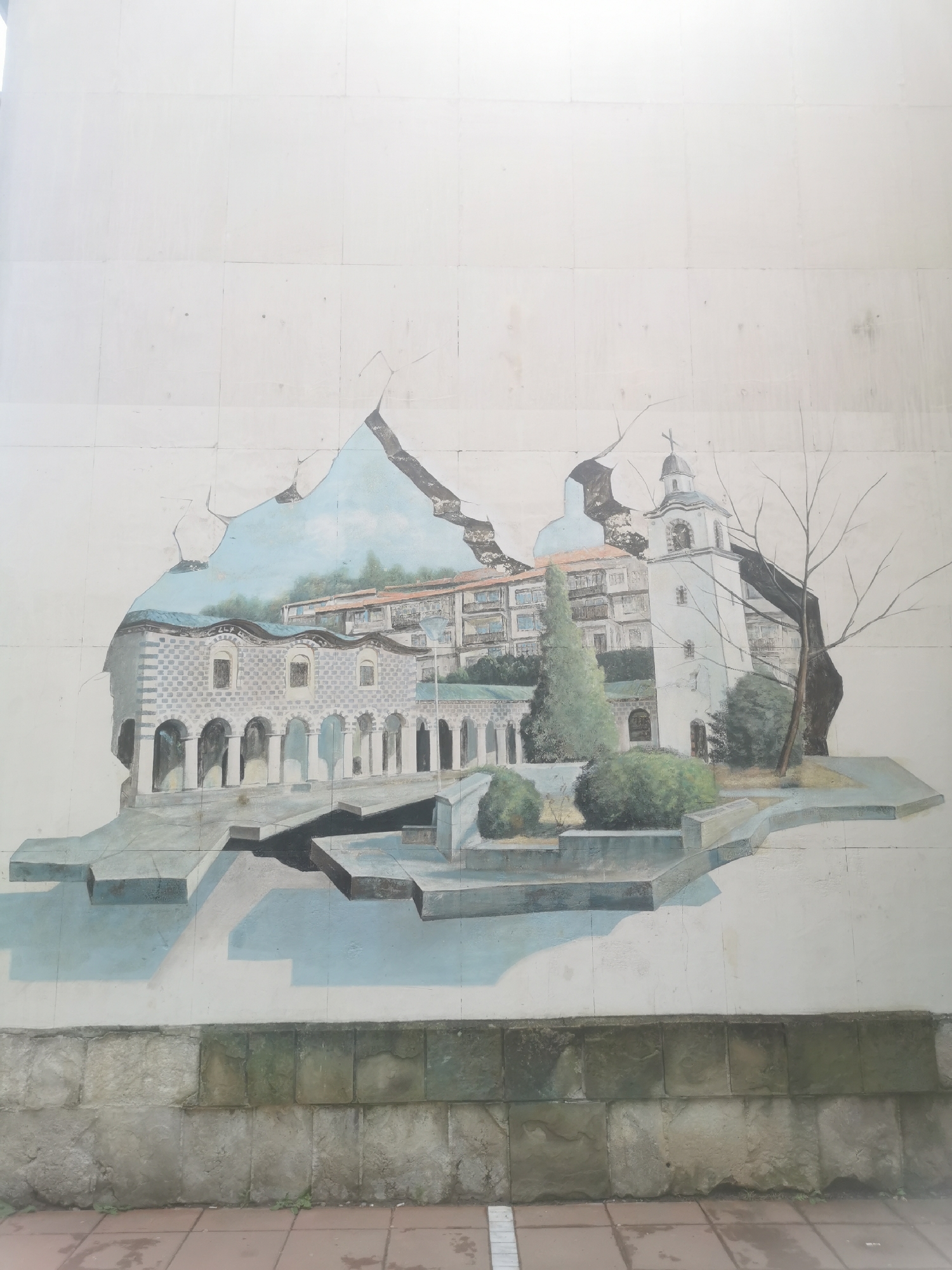 A large outdoor mural is painted on a light-colored, tiled wall, creating a trompe l'oeil effect as if a scene is breaking through the surface. The mural depicts an architectural landscape, likely a cityscape.
On the left, a stone building with an arched colonnade or gallery is visible, featuring dark windows and a distinctive roofline. Behind it, a multi-story building with numerous windows and brown-tiled roofs appears, nestled against a backdrop of blue sky and distant hills or mountains. To the right, a prominent white church or bell tower with a domed top and a cross stands tall, flanked by a bare, deciduous tree and several green bushes and a tall coniferous tree. The ground within the mural is rendered with steps and pathways, casting shadows that enhance the illusion of depth.
Below the painted scene, the actual wall transitions into a rough, rectangular stone base, suggesting a foundation or lower part of the structure. The very bottom of the image shows reddish-brown tiled pavement.
The scene is illuminated as if by daylight, with soft shadows indicating a light source from the left, highlighting the dimensional quality of the painting. No human figures or discernible activities are depicted, focusing solely on the architectural and natural elements. There is no visible text in the image. The location is Blagoevgrad, Bulgaria.