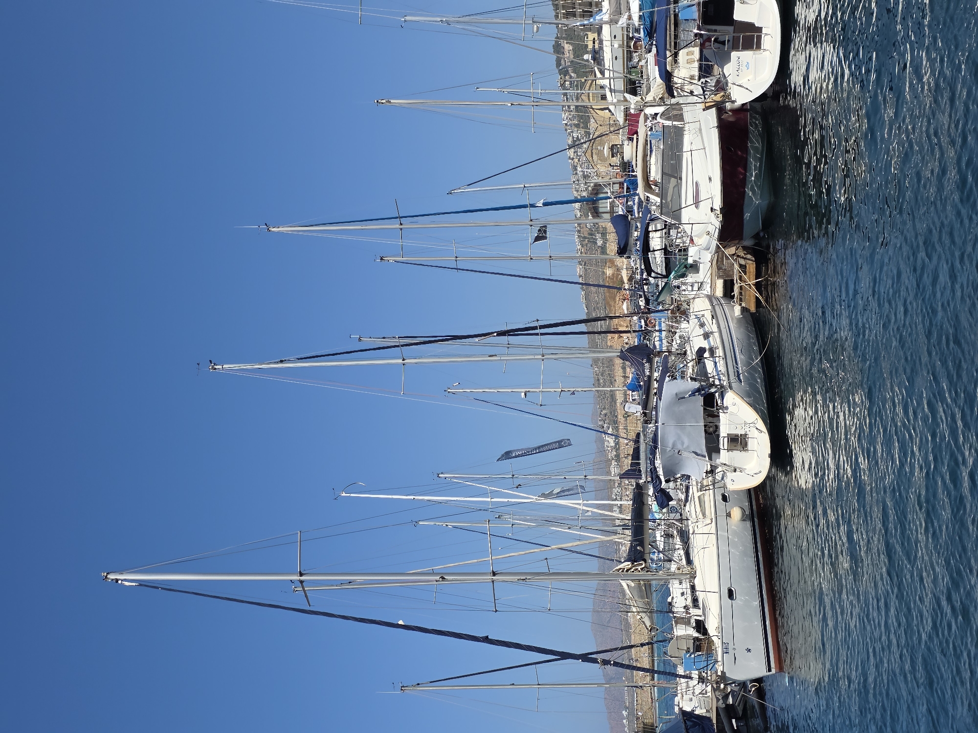 The image shows a marina filled with numerous sailboats and a few motorboats docked in calm, blue water under a clear, bright blue sky. The boats are moored in rows, with their tall masts dominating the scene and casting shadows on the water. In the background, a hillside covered with buildings and vegetation slopes down towards the harbor. The architecture suggests a Mediterranean setting, and the presence of a flag with text, possibly in Greek, further supports this. The water is slightly rippled, reflecting the sunlight and the surrounding boats. The overall atmosphere is one of a peaceful, sunny day at a harbor, likely in a coastal town. Given the architecture and the clear Mediterranean blue sky and water, this scene is consistent with the port of Chania, Crete, Greece, known for its Venetian harbor and active marina.