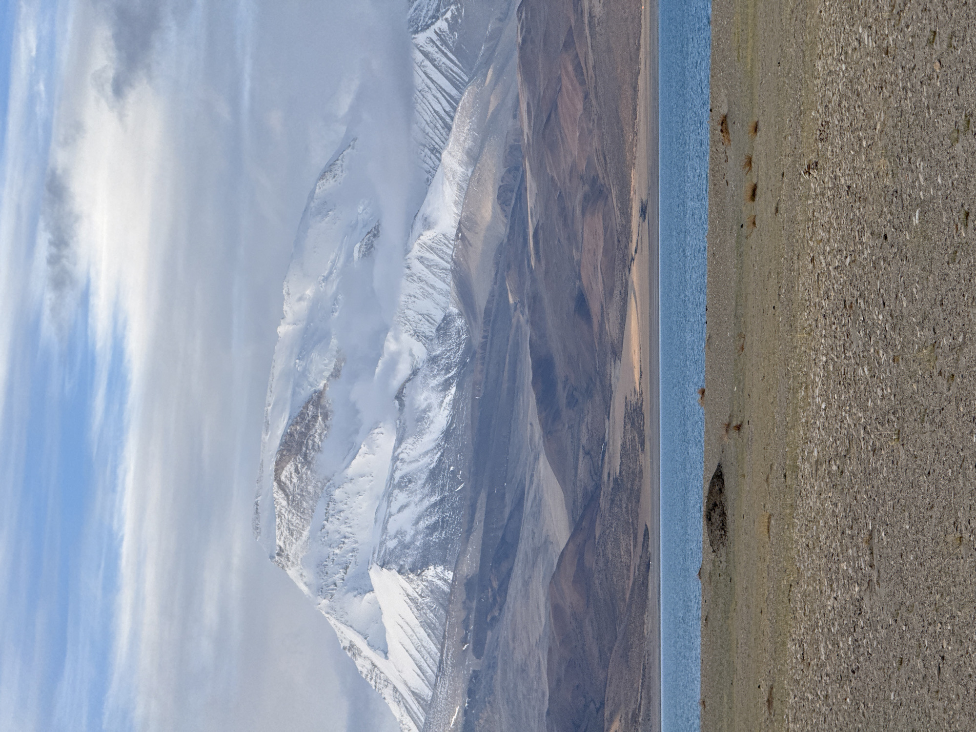 This image displays a mountainous landscape next to a large body of water, likely a lake. The foreground shows a rocky, barren shoreline transitioning to the lake's blue surface. Beyond the lake, imposing mountains rise, some covered with snow and ice, partially obscured by clouds. The image was taken in Purang, Tibet, China. The left side of the image has a sliver of blue sky with wispy clouds.