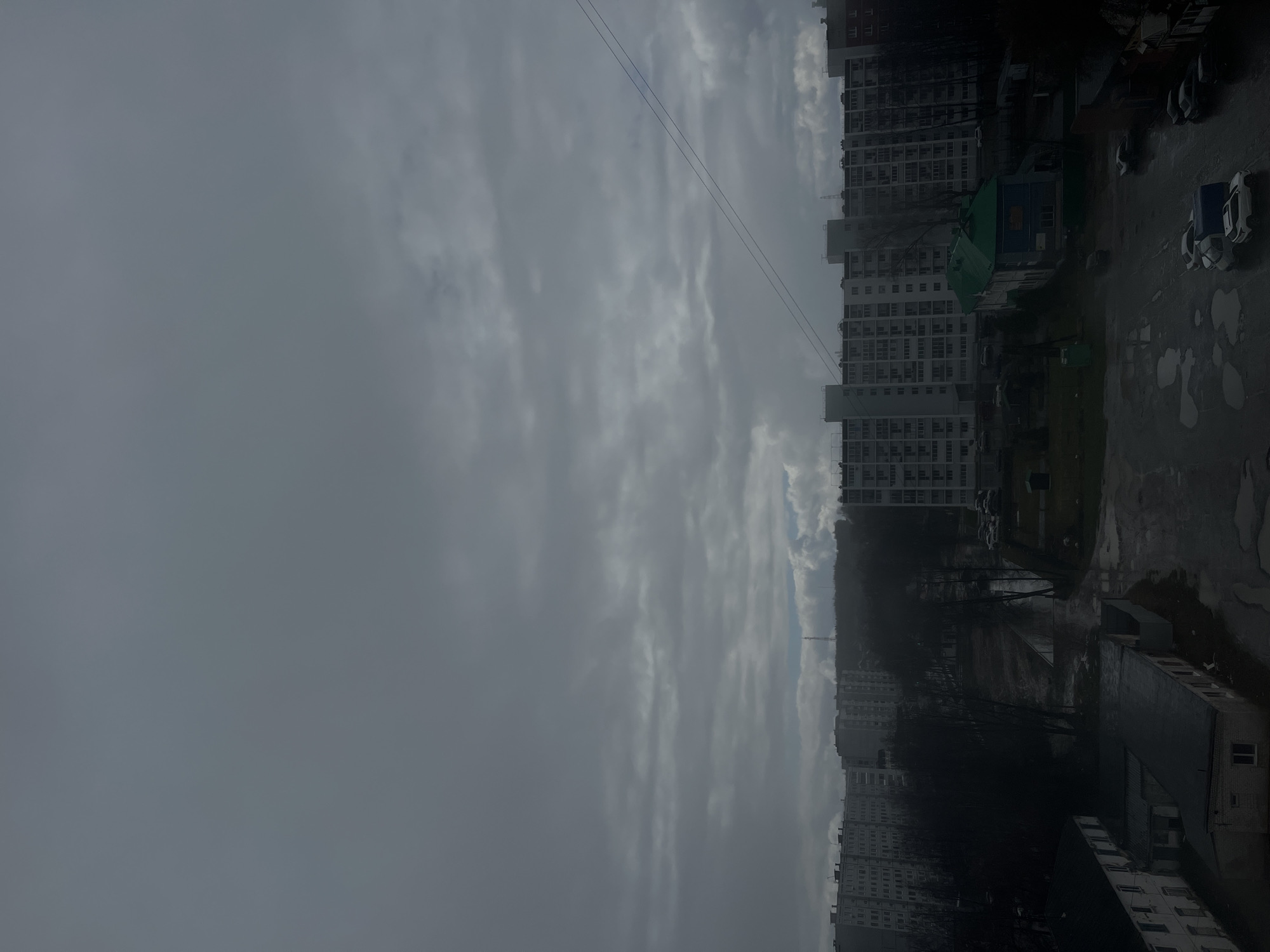 The image shows an urban landscape in Chaikovsky, Russia, under a cloudy sky. Tall apartment buildings line the right side of the frame. A small green building stands out among the larger structures. Below the buildings, a paved area shows puddles and parked cars. Power lines are visible across the sky.
