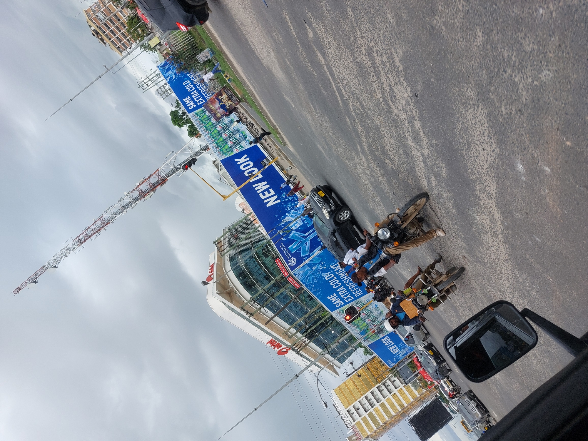 The image appears to be taken from inside a vehicle in Dar es Salaam, Tanzania. The scene is dominated by a busy streetscape. In the foreground, the side mirror of the vehicle is visible. Adjacent to the vehicle is a series of motorbikes, some occupied by riders. A black car and other vehicles are also present on the road. Large advertising banners displaying the phrases "NEW LOOK" and "SAME EXTRA COLD REFRESHMENT" in blue are prominently featured alongside images of bottled beverages. These banners are affixed to building structures, including one that appears to be under construction with a tall crane extending into the sky. Buildings of different architectural styles are visible, including modern glass-paneled structures and older buildings with gold colored decorations. The sky is overcast with grey clouds.