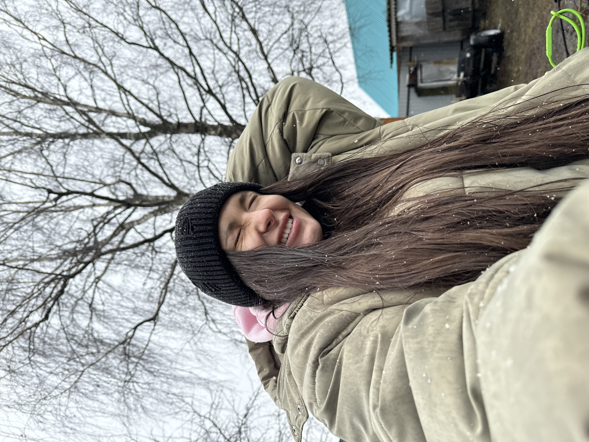 A young woman with long brown hair and a black beanie is smiling widely at the camera. She is wearing a beige puffer jacket, and there are snowflakes on her hair and jacket. In the background, there are bare trees and a building with a turquoise roof. The location is Unknown, Unknown.