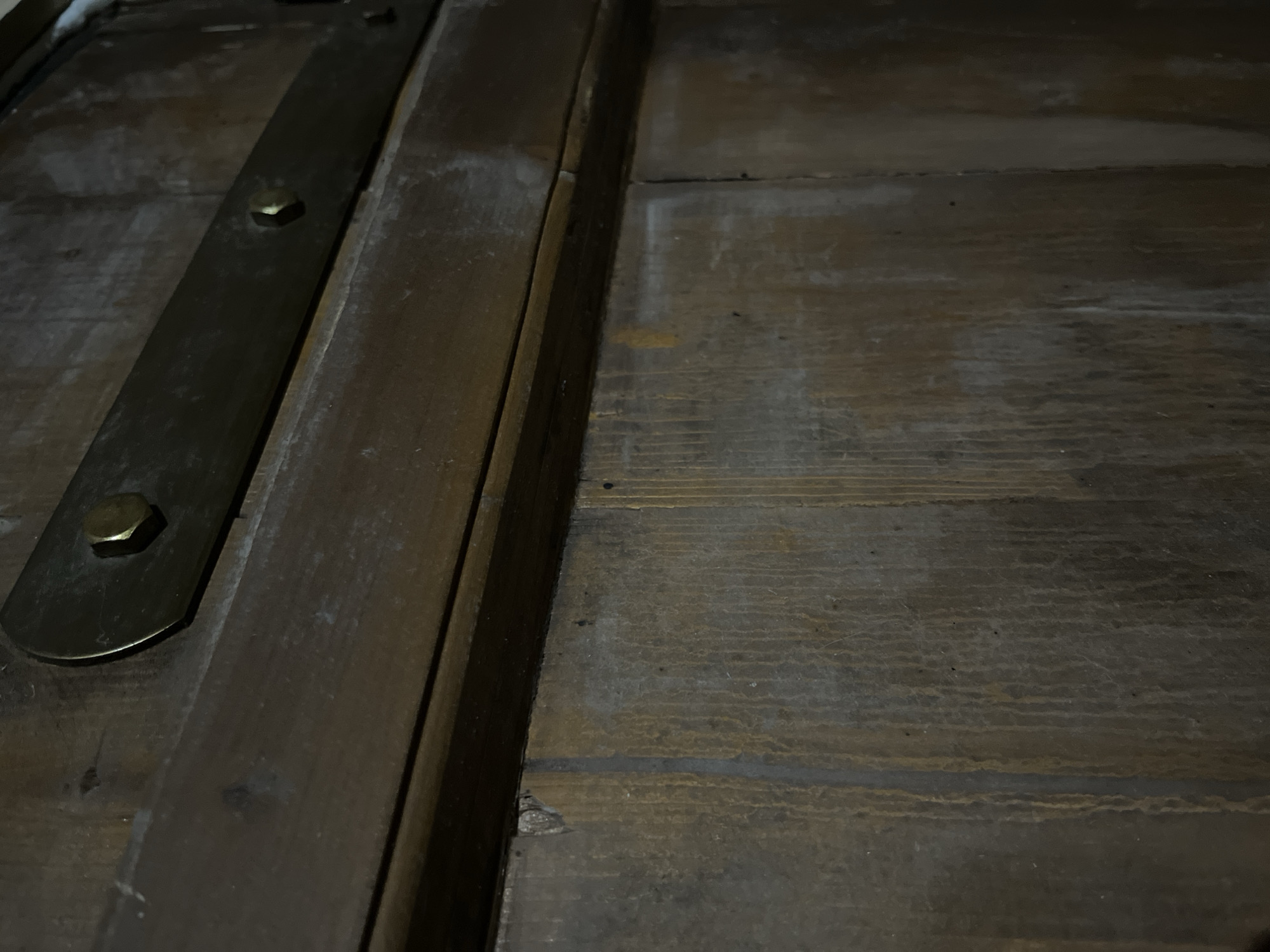 The image shows a close-up view of a wooden surface, possibly the top of an antique chest or piece of furniture. The wood is stained in a dark brown color and exhibits a textured grain pattern. A metal strap or brace runs vertically along the left side, affixed with hexagonal bolts. The overall environment appears dimly lit, contributing to a somber tone. There are no people, actions, or interactions visible in the frame.