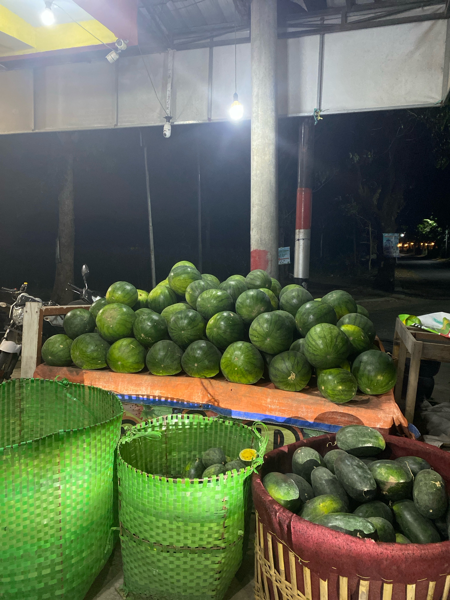 The image depicts a nighttime outdoor scene at what appears to be a fruit stand in Kediri City, Indonesia. A large pile of watermelons is prominently displayed on a flat surface covered with an orange tarp. The watermelons are round and have a dark green striped rind. To the left of the watermelon pile, there are two large, woven green baskets, one of which contains a few watermelons. To the right of the watermelon pile, there is a woven basket filled with elongated, dark green fruits, possibly also watermelons or a similar variety. In the background, a motorcycle is parked on the left, and beyond that, a street with some trees and distant lights is visible. The scene is illuminated by artificial lighting, including a bare light bulb hanging from a structure above. There are no people visible in the image. The environment appears to be urban, with a paved road and some structural elements suggesting a covered market or roadside stall.
