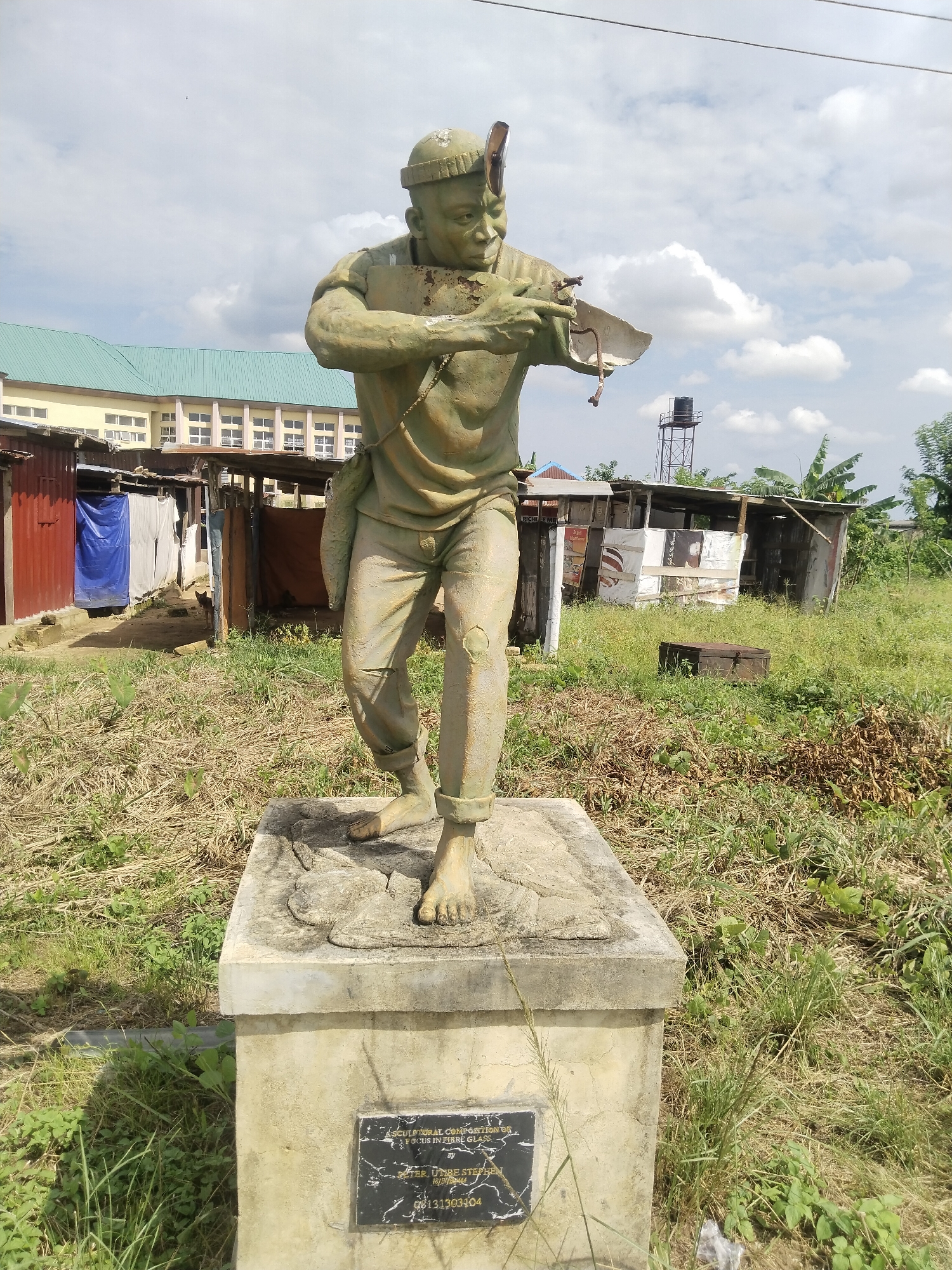 The image depicts a green, weathered statue of a man standing on a concrete pedestal. The man is barefoot, wearing pants and a shirt, and has a tool in his hands. The statue is set in an overgrown grassy area with dilapidated shacks in the background. A water tower is visible behind the shacks. A building with a green roof is visible further in the background. The statue's pedestal has a plaque with the text "A SCULPTURAL COMPOSITION FOCUS IN FIBRE GLASS BY PETER, UMORE STEPHEN 18/07/2012 08131303404." The statue is located in the city of Uyo, Nigeria.