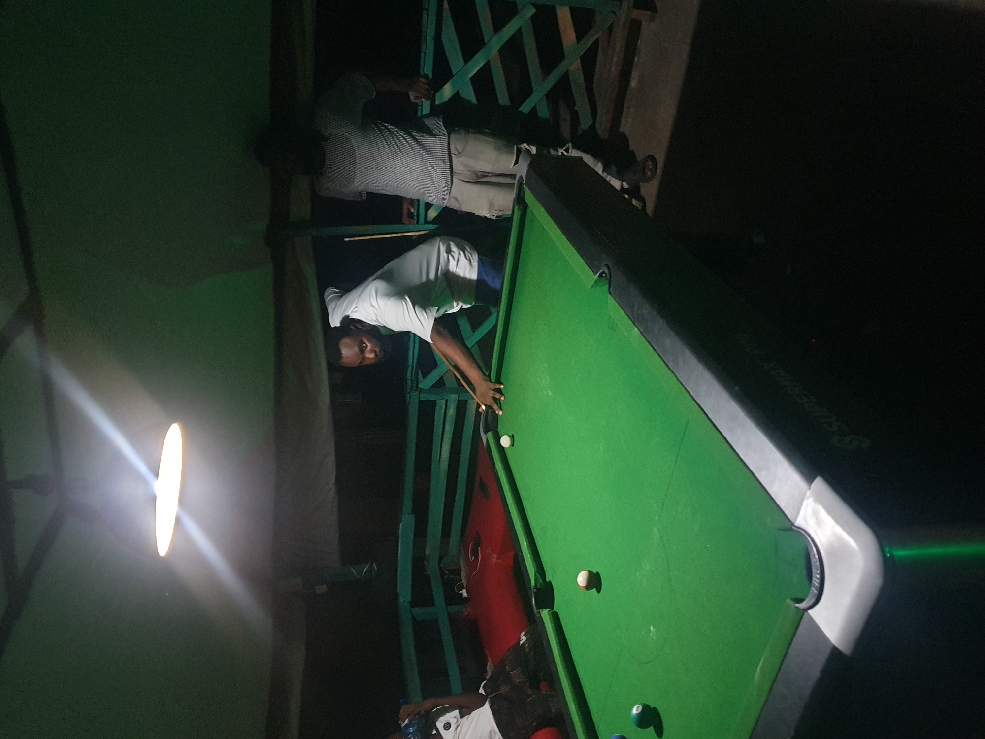 In Lokoja, Nigeria, a group of men are engaged in a game of billiards. The scene is dimly lit, with a bright overhead light illuminating the pool table. One man, wearing a white t-shirt and shorts, is actively playing, his body bent over the table, cue stick in hand. Another man, in a checkered shirt and torn pants, stands nearby, observing the game. A third individual, partially visible, is seated in the background. The pool table, green with white balls scattered across its surface, is the central focus of the image. The environment appears to be an outdoor or semi-outdoor space, possibly a bar or recreational area, with a wooden railing and dark background suggesting the absence of significant city lights. The overall atmosphere suggests a casual and relaxed gathering.