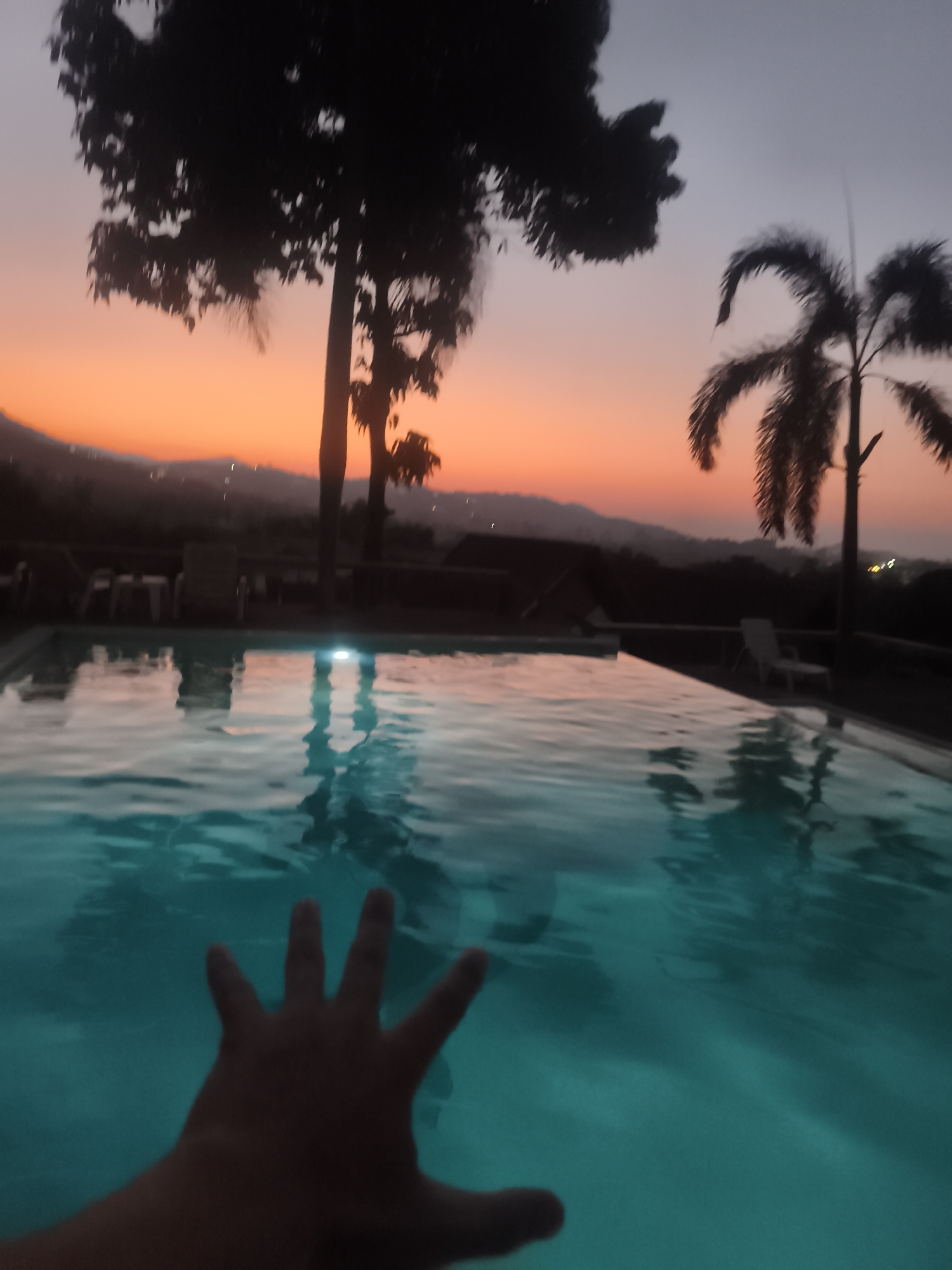 A person's hand is partially submerged in a turquoise swimming pool, reflecting the twilight sky. The background features silhouetted trees and palm trees against an orange and purple sunset. A distant mountain range is visible. Chairs and tables are situated near the pool's edge. The location is likely in or near บ้านบางรักษ์, Thailand.