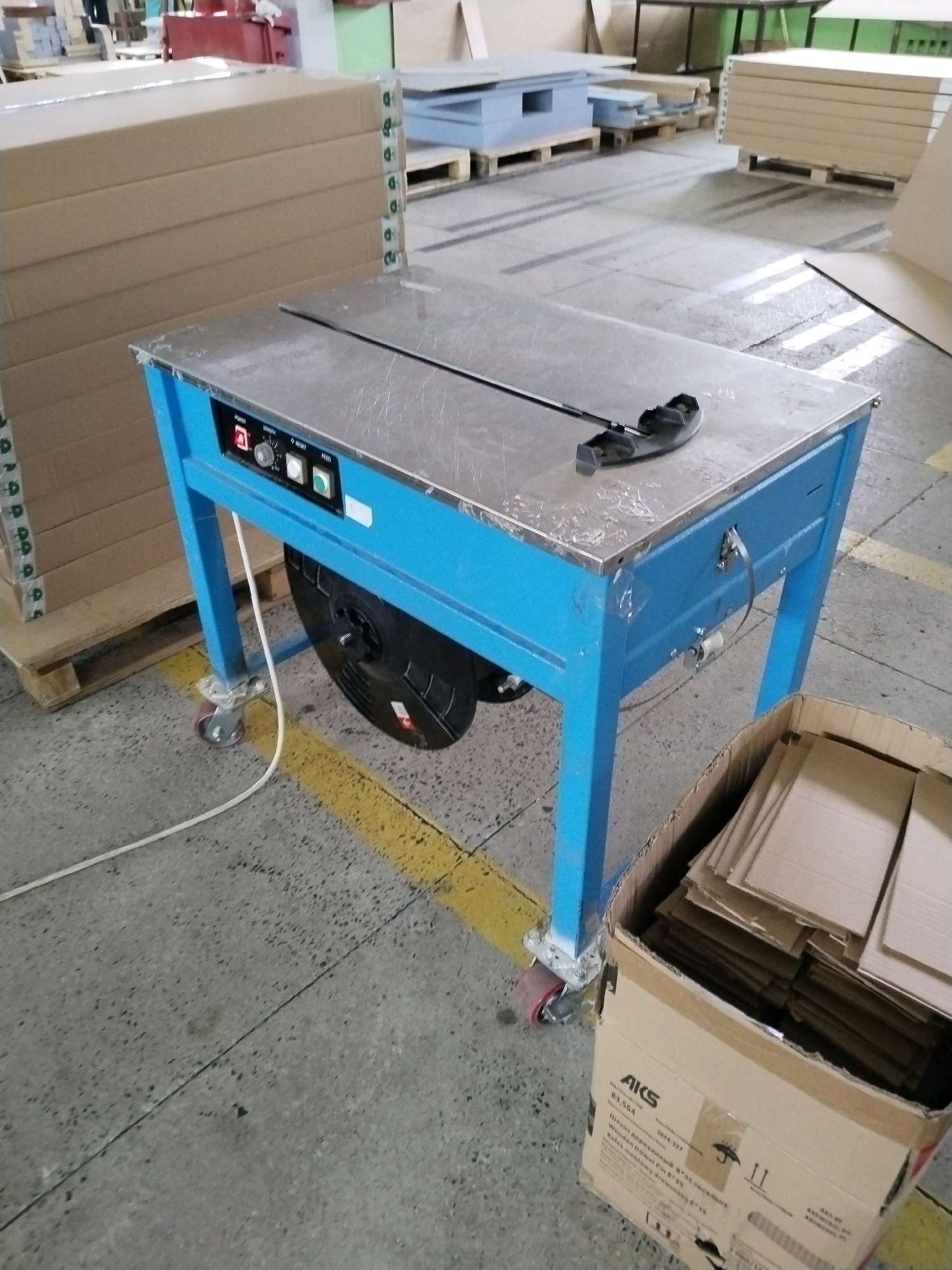 In a warehouse setting within the city of Гожскі сельскі Савет, Belarus, a blue strapping machine is centrally positioned. The machine features a silver-colored steel surface, control buttons labeled "POWER," "TENSION," and "RESET," and a red power indicator light. A black strapping reel is attached to the underside of the machine. To the left of the strapping machine, stacks of large cardboard boxes are visible, indicating packaging or shipping materials. In the foreground to the right, a cardboard box filled with smaller cardboard pieces is present. The floor is concrete, marked with yellow lines. The background shows additional shelving units, pallets, and more cardboard materials, suggesting an active industrial or storage environment. No people are present in the image.