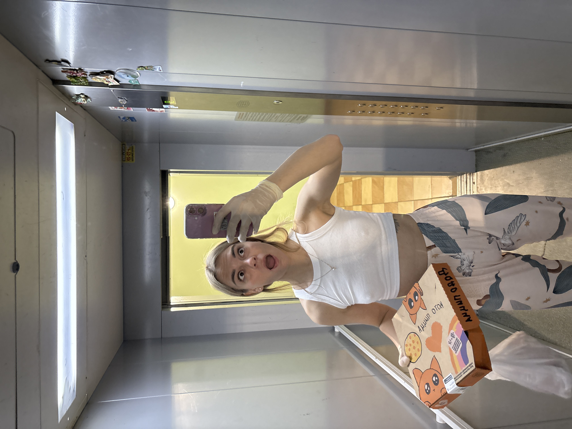 A young woman is captured in a mirror selfie inside an elevator. She has blonde hair and a surprised expression, with her mouth agape. She wears a white ribbed crop top, patterned pajama pants adorned with bird and wave designs, and a transparent glove on her left hand. This hand holds a smartphone with a purple or pink case, pointed towards the mirror for the selfie.
In her right hand, she carries a cardboard box decorated with cartoon cats, a pizza slice, a heart, and a rainbow-colored ribbon motif. Visible text on the box includes "BOBO LINKY", "АМНИТ ОГОН" (likely part of the Russian slang "АМНИТ ОГОНЬ"), and "91-16".
The elevator features metallic walls and a large mirror on the right side. Through the open elevator door, a bright yellow wall and light-colored tiled floor of a hallway are visible. Fluorescent lighting illuminates the elevator. Small magnets or decorative items are affixed to the metal frame above the door. The setting is an indoor location, consistent with an elevator in a building in Krasnoyarsk, Russia. The woman's expression suggests a playful or humorous mood.