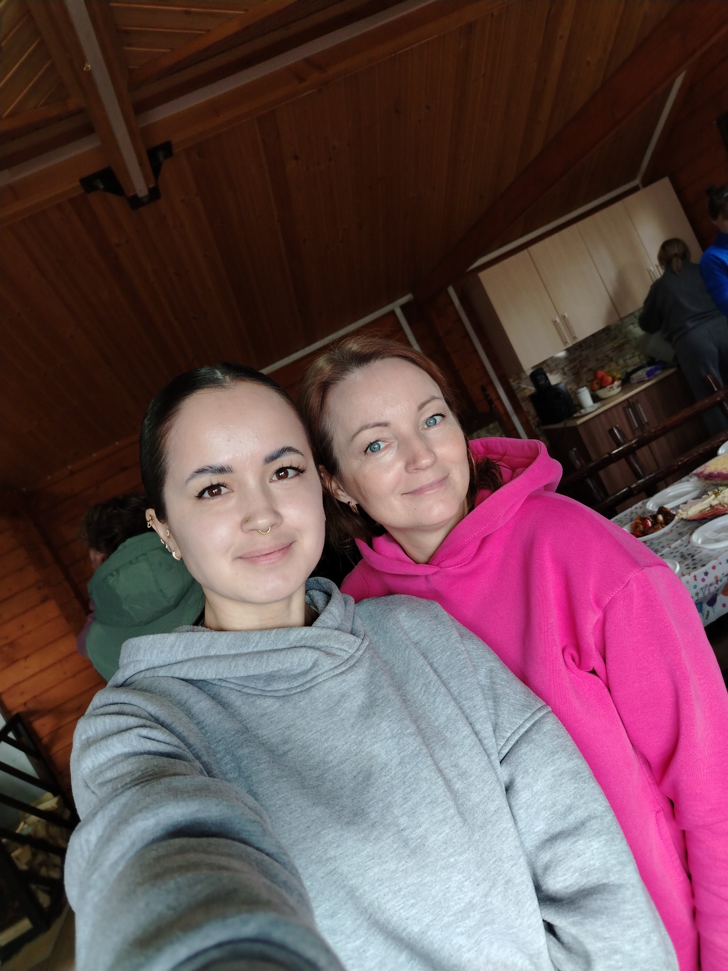The image depicts two women in an indoor setting, likely a wooden cabin. The woman on the left is wearing a grey hooded sweatshirt and has dark hair pulled back. She has a nose ring and multiple earrings. The woman on the right is wearing a bright pink hooded sweatshirt and has reddish hair. In the background, other people are visible near a kitchen area with cabinets and a table. A table with food items is in the lower right. The ceiling and walls are wood paneled. The location is potentially in the city of Chaikovsky, Russia, based on the user's request.