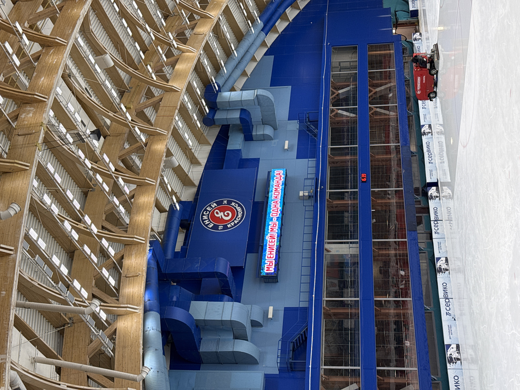 This image captures a large indoor ice arena, likely viewed from an elevated position. The scene is dominated by a complex, light-colored wooden lattice ceiling structure on the left, featuring numerous bright white lights arranged in rows. Adjacent to this structure on the right, a multi-story wall in shades of blue and grey houses industrial-style ventilation ducts and panels, with large windows providing a glimpse into interior spaces.
At the bottom right, a pristine white ice rink is visible, upon which a bright red ice resurfacer (similar to a Zamboni) is operating, suggesting ice maintenance or preparation. The resurfacer bears the text "черноголовка" (Chernogolovka, a Russian beverage company) and "OLYMPIA", indicating its brand. Along the edge of the ice rink, multiple white banners display logos and text, including "сервико" (Serviko), "ЛОГИСТИКА" (Logistics), and "СИБЭНКО" (SibEnko), likely sponsors. A digital clock on the blue wall shows "16:39".
Prominently displayed on a blue wall section is a large red circular logo featuring the text "ЕНИСЕЙ" (Yenisei), "19", "34", and "КРАСНОЯРСК" (Krasnoyarsk), strongly indicating the venue is home to a team or organization named Yenisei from Krasnoyarsk, Russia. Below this, an illuminated blue sign reads "МЫ ЕНИСЕЙ! МЫ - ОДНА КОМАНДА!" (We are Yenisei! We are one team!), reinforcing the team's identity. The overall impression is of a modern, well-equipped sports facility in Krasnoyarsk, geared for ice-based sports.