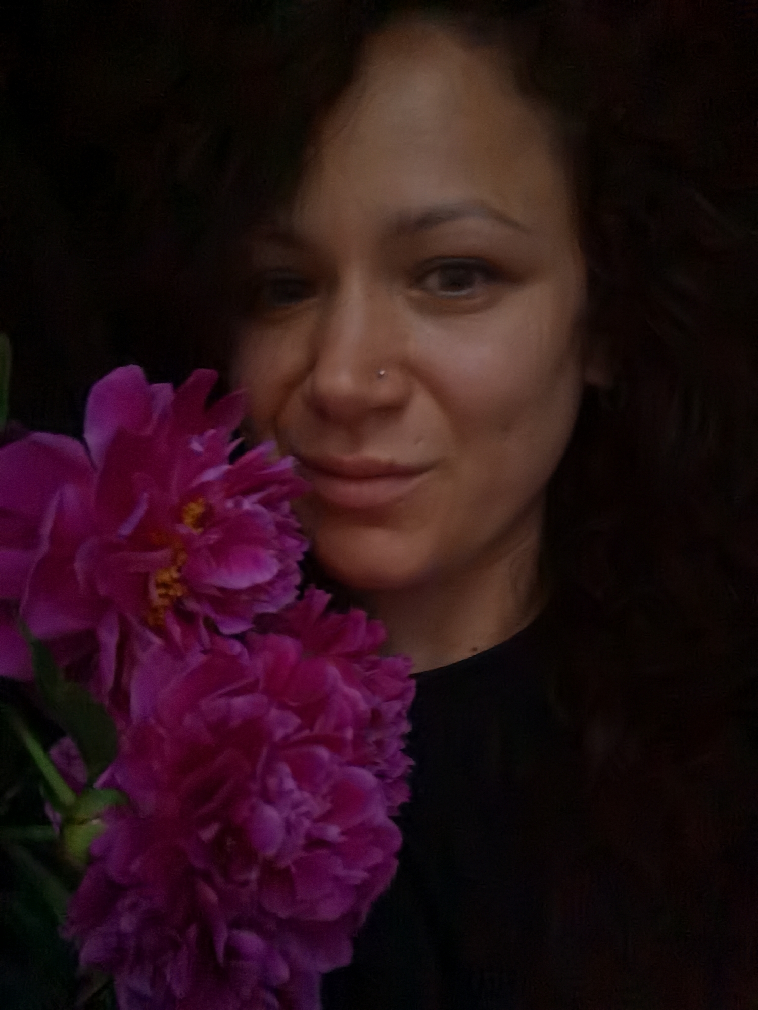A woman with dark curly hair and a nose ring is holding a bouquet of bright pink peonies in front of her face. She is wearing a black shirt. The background is dark and out of focus. The image appears to be a selfie taken indoors. There is no information in the image to determine its location in Chaikovsky, Russia.
