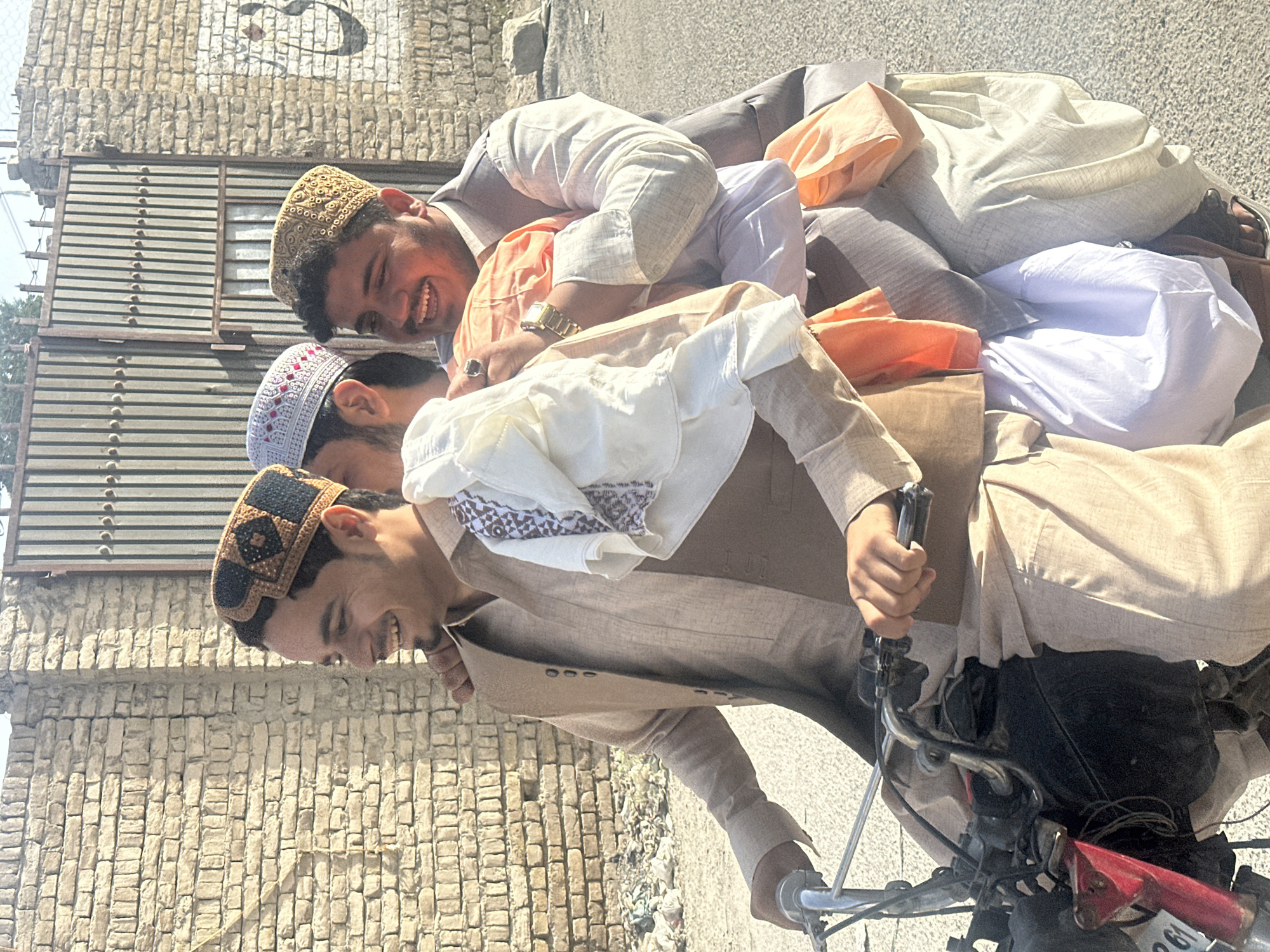A group of four young men are riding together on a motorcycle on an asphalt road in Quetta City Tehsil, Pakistan. They are all dressed in traditional Pakistani clothing, including caps on their heads. The rider is wearing a brown outfit, while the others are wearing various light-colored outfits. The motorcycle is red and black. A brick building and metal structure are visible in the background.