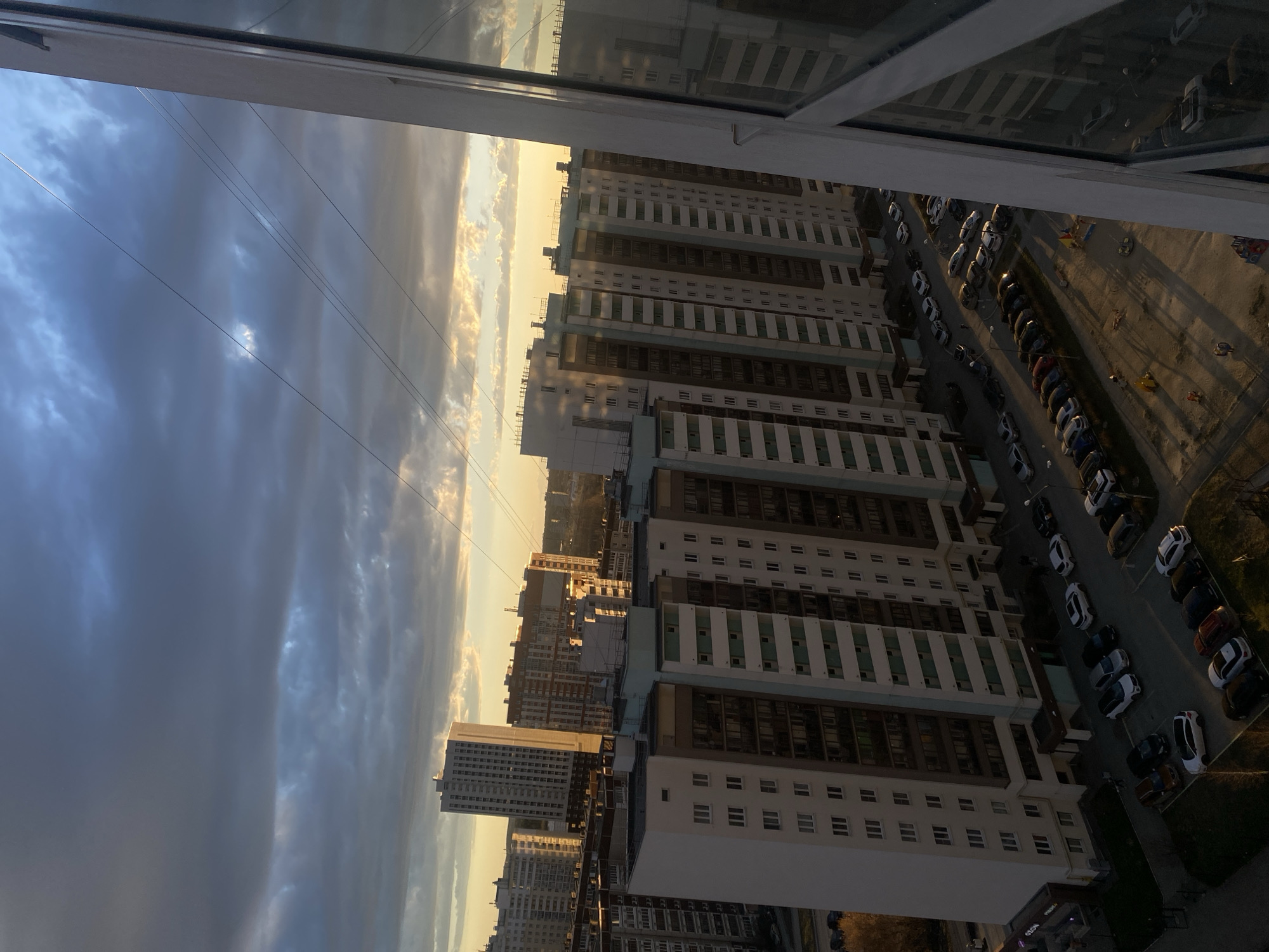 This image presents a high-angle view of an urban residential area, likely captured from a window, as indicated by the window frame visible at the top. The scene depicts a sprawling cityscape under a partially cloudy sky, illuminated by the warm, golden light of a low-hanging sun, suggesting late afternoon or early evening, possibly sunset.
The landscape is dominated by numerous multi-story apartment buildings, characteristic of modern residential complexes in cities like Yekaterinburg, Russia. These buildings feature contemporary architecture, with facades predominantly in light colors (white or light grey) accented by darker windows and occasional light blue or green panels. The sun casts long shadows from the buildings across the ground.
Below the buildings, a wide paved street or parking area stretches out, densely lined with a variety of parked passenger cars. Adjacent to this, a small playground with colorful equipment, including what appears to be a swing set, is discernible, although no people are visible in the scene. Several thin, parallel wires, likely utility lines, crisscross the sky in the left portion of the image.
No human activity or individuals are explicitly depicted. The overall mood is quiet and contemplative, capturing the scale of urban development at dusk. On a sign at the base of one of the buildings, faint text resembling "VISTO" or "VOSTO" can be partially seen.