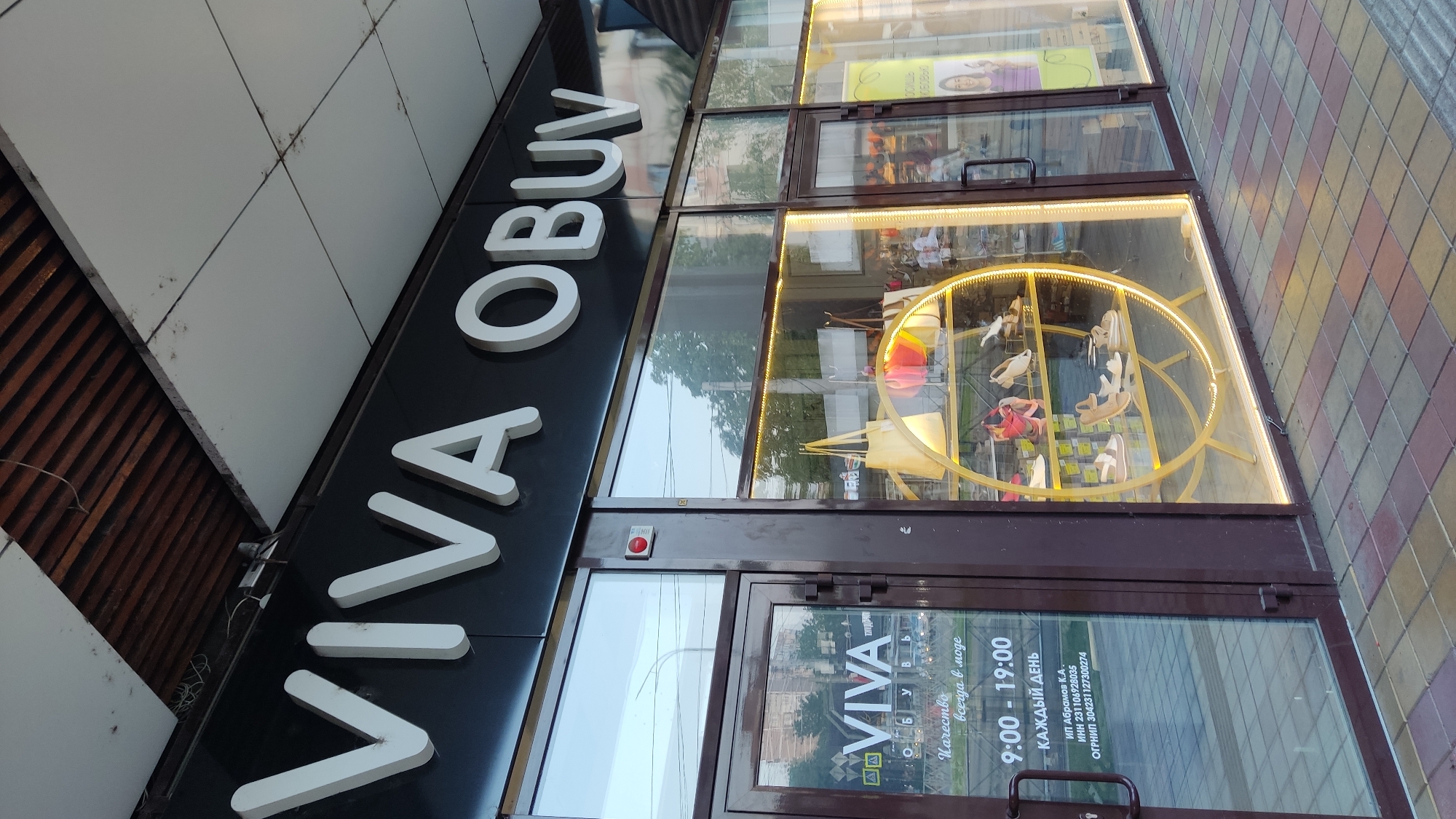 The image shows the storefront of a shoe store called "VIVA OBUV" in Krasnodar, Russia. The store features a large, white, three-dimensional sign with the name "VIVA OBUV" above the entrance. The storefront has large glass windows displaying shoes and handbags on a circular gold shelf. A door with "VIVA" and business hours printed on it is visible to the left. The exterior of the building is dark brown with white trim, and the pavement in front of the store is made of colorful square tiles. The lighting suggests it is daytime.