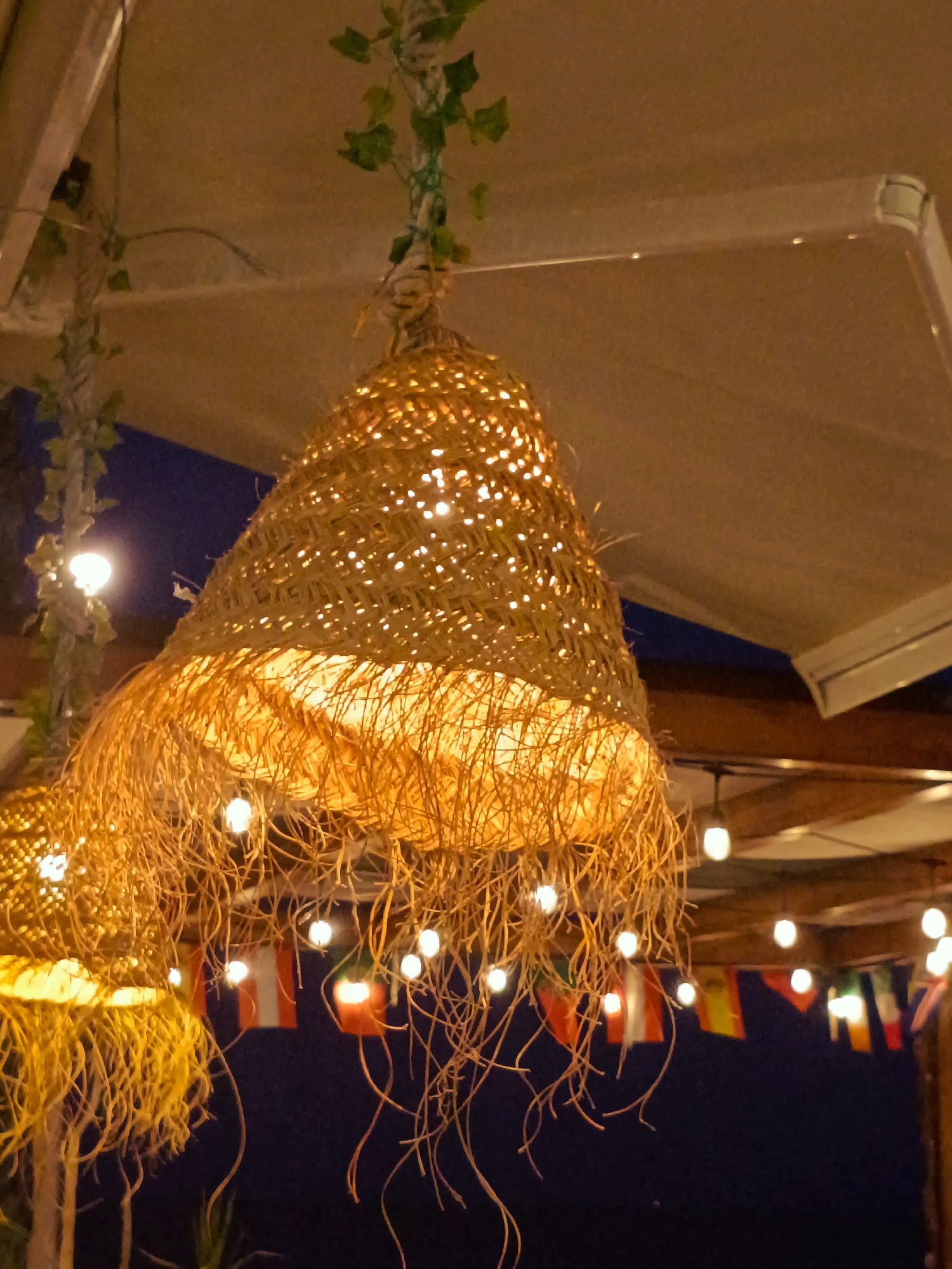 This image captures an outdoor dining or relaxation area at night, likely a terrace or patio, with a focus on decorative lighting and ambiance. The foreground is dominated by two large, conical lampshades made of woven straw or dried grass. These lampshades are illuminated from within, casting a warm, golden glow and revealing their intricate weave. Dangling from the bottom of these lampshades are numerous strands of the same straw-like material, creating a fringe effect. Small, white string lights are also visible, some integrated with the lampshades and others strung along a wooden beam in the background.
Behind the lampshades, a dark blue night sky is visible. Across the background, a string of small flags, in various colors including red, white, yellow, and green, is hung. These flags appear to be decorative and do not clearly represent any specific national or organizational insignia. The area is covered by a light-colored awning or canopy. The overall impression is one of a relaxed, possibly coastal or Mediterranean, setting, typical of a restaurant, bar, or resort. No people are visible in the image. The setting could be San Gabriel, Spain, a municipality in the province of Alicante, known for its coastal areas and tourist amenities.