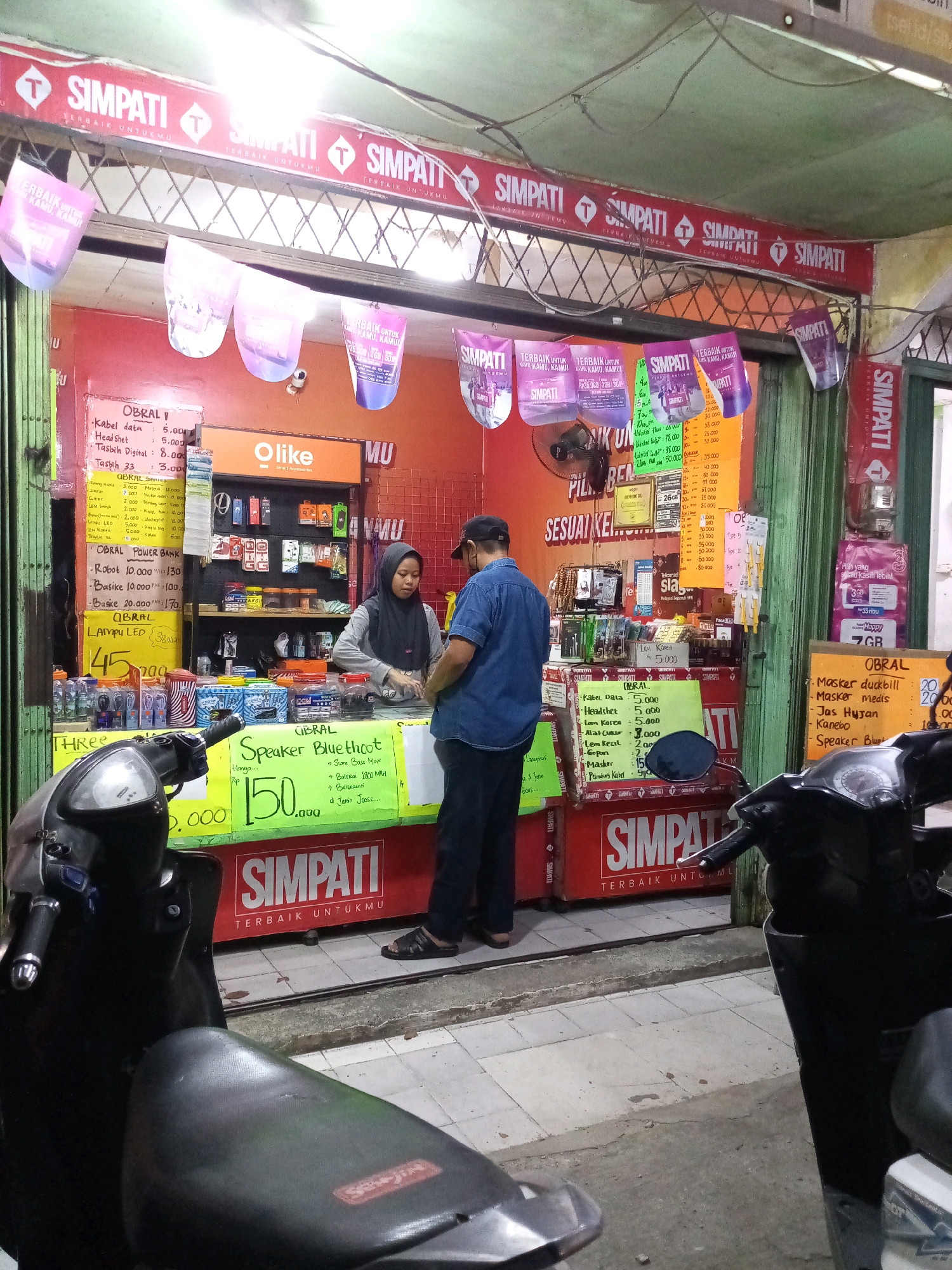 The image captures a street-level view of a SIMPATI telecommunications store in Bekasi, Indonesia, at dusk. The store features a red facade with prominent SIMPATI branding and an overhang adorned with promotional banners. A woman wearing a hijab and a man in a denim shirt are seen interacting near the counter. Various accessories like cables, headphones, and power banks are displayed on shelves and in promotional signs. A motorbike is parked in the foreground, partially obscuring the store entrance. The pavement in front of the store is tiled, and a slightly worn red store sign is visible. The overall scene suggests a bustling retail environment in a local urban setting.