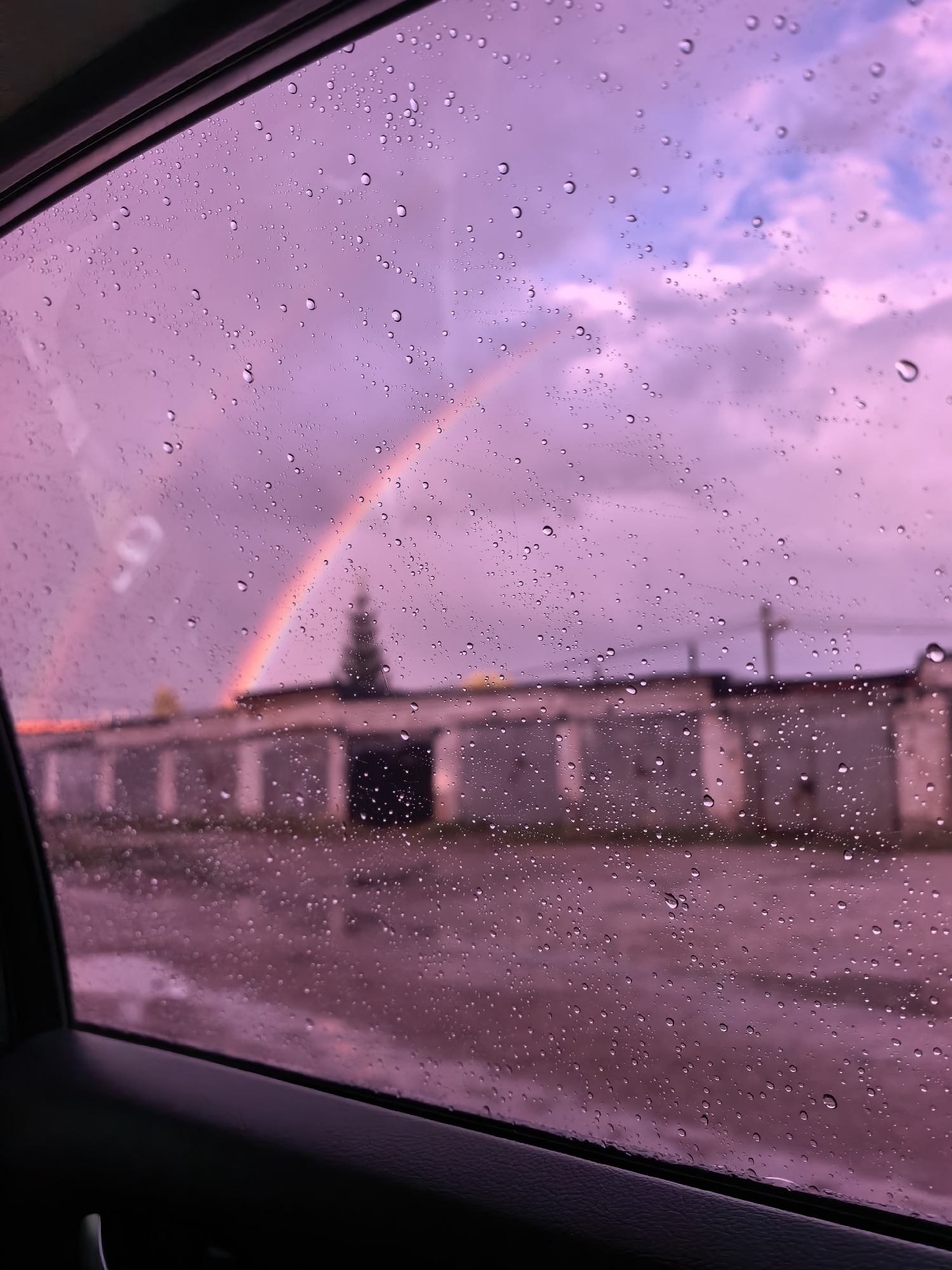 The image depicts a view through a car window on a rainy day, with a rainbow visible in the sky. The window is covered in water droplets, distorting the view of the outside. In the background, there is a long, low building that appears to be a row of garages or storage units, typical of urban areas in Russia. A dark silhouette of a tree is visible behind the building, to the left of the rainbow. The sky is a mix of purple and pink hues, suggesting either dawn or dusk, with some clouds. The ground in front of the building appears wet and reflective. The composition suggests the photo was taken from inside a car, with the dark interior of the car door visible in the foreground on the left. The image does not show any people. The location is identified as Sterlitamak, Russia.