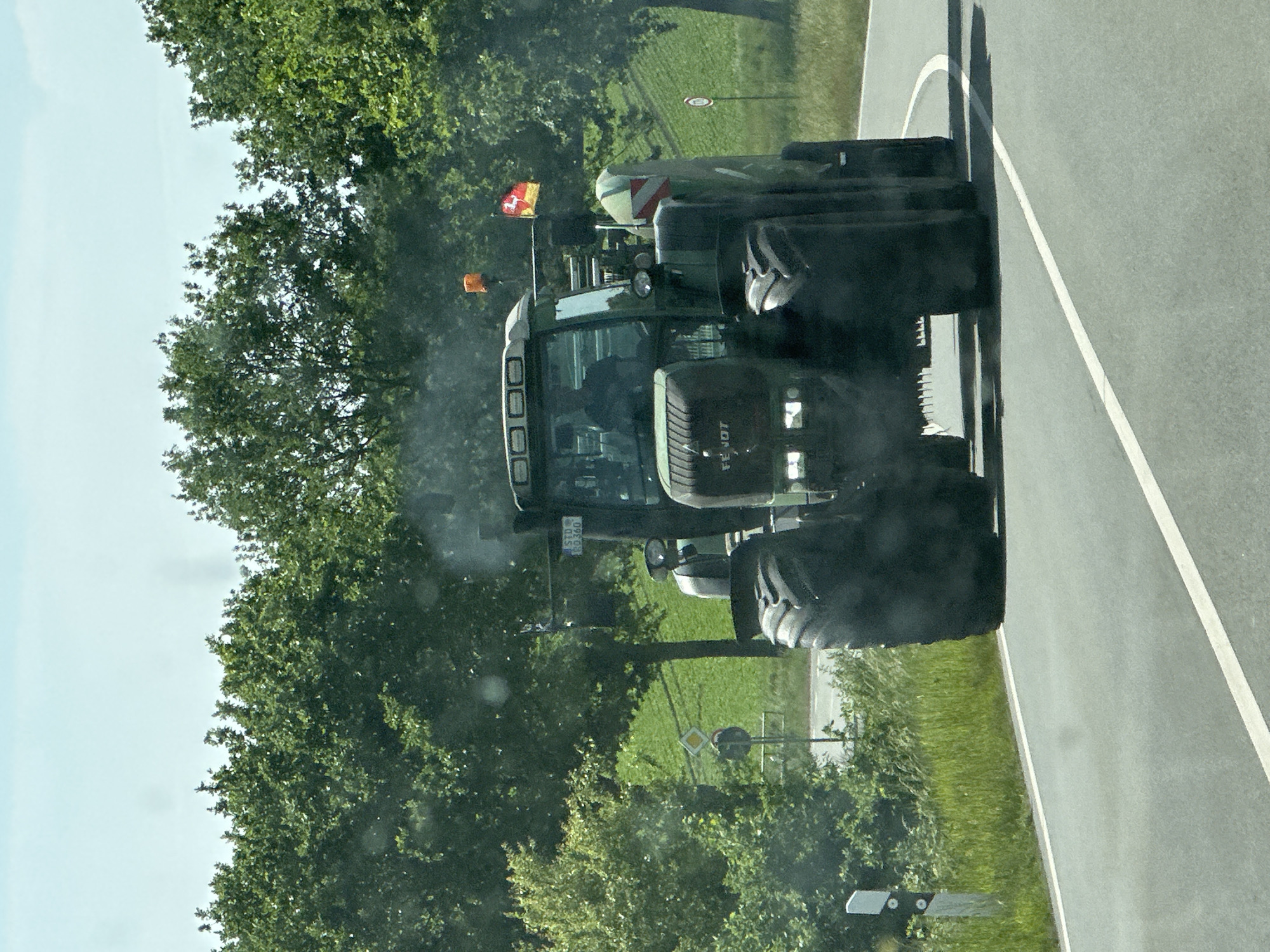 A Fendt tractor is on the road in Buxtehude, Germany, alongside a tree-lined field. The tractor is traveling on an asphalt road with a white line, indicating lanes. The tractor is green and has the Fendt logo on the front. It is equipped with large tires. A tree-lined field borders the road, with green grass and various road signs visible. The sky is partially visible, suggesting daylight conditions. The tractor has a license plate that reads "STD RD 360."