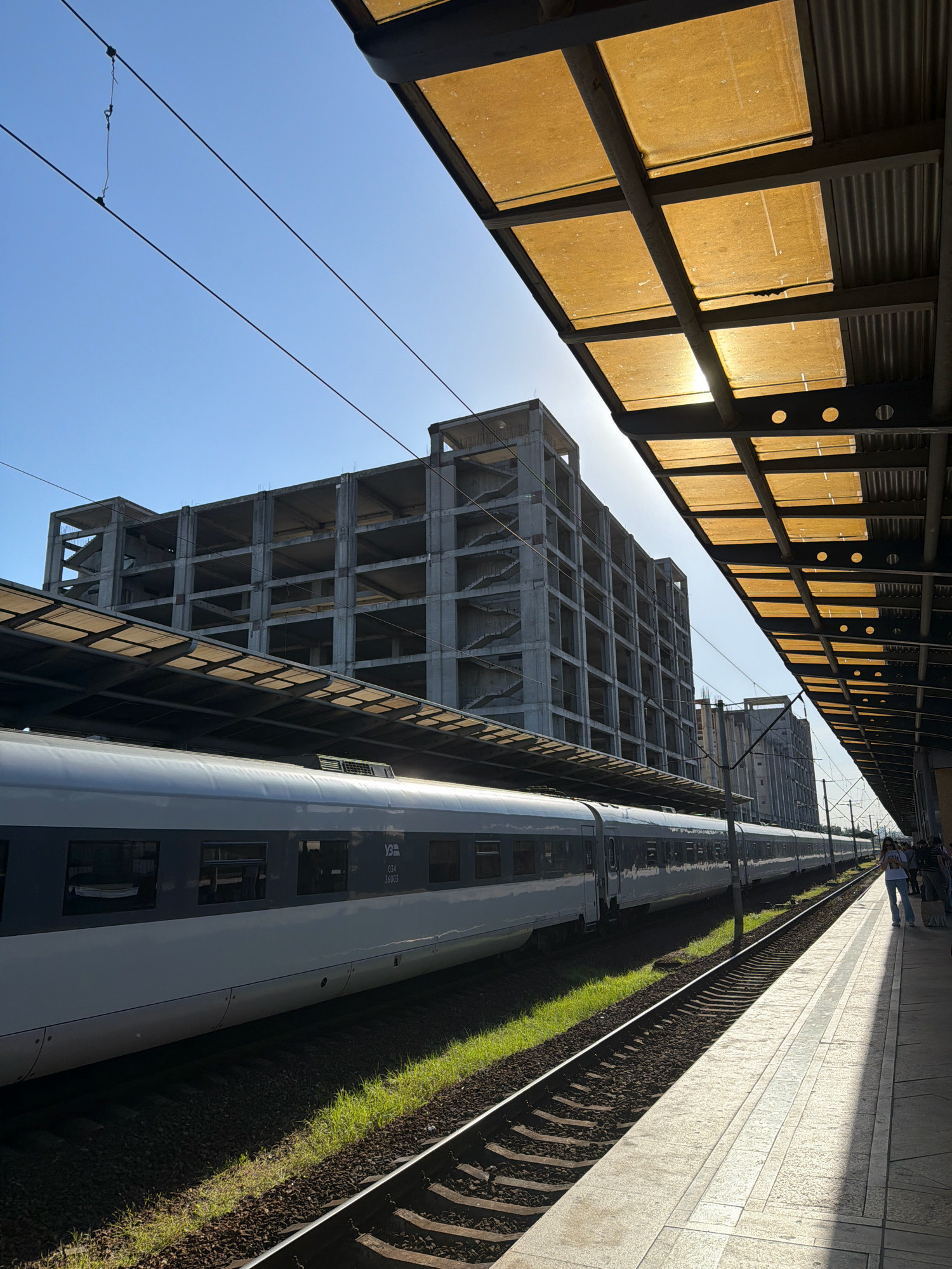 This image captures a train at a Kyiv, Ukraine railway station, likely Kyiv-Pasazhyrskyi, given the architecture and infrastructure visible. The scene is bathed in bright sunlight, creating distinct shadows. In the foreground, a long, sleek, modern train with white and dark grey carriages is stopped at the platform. The train is identified with markings such as "034 36003" and a symbol that appears to be "Укрзалізниця," the Ukrainian Railways logo.
Adjacent to the train is a concrete platform with patterned tiles, some of which are illuminated by the sun. Overhanging the platform is a canopy structure supported by metal beams, featuring translucent yellow panels that allow sunlight to filter through, creating a warm glow. In the background, a large, unfinished concrete building dominates the skyline. This multi-story structure, with its exposed rebar and open floors, suggests ongoing construction or a long-abandoned project. Further in the distance, other buildings, possibly industrial or utility structures, are visible.
A few people are present on the platform, including a woman in a white t-shirt and jeans, standing and looking at her phone. Another person, wearing a backpack and dark clothing, is also visible further down the platform. The sky is a clear, bright blue, indicating a sunny day. The railway tracks are clearly defined, with ballast and sleepers visible. The presence of overhead electric wires confirms this is an electrified railway line. The overall impression is one of urban infrastructure, with a contrast between modern transportation, ongoing development, and the bright natural light.