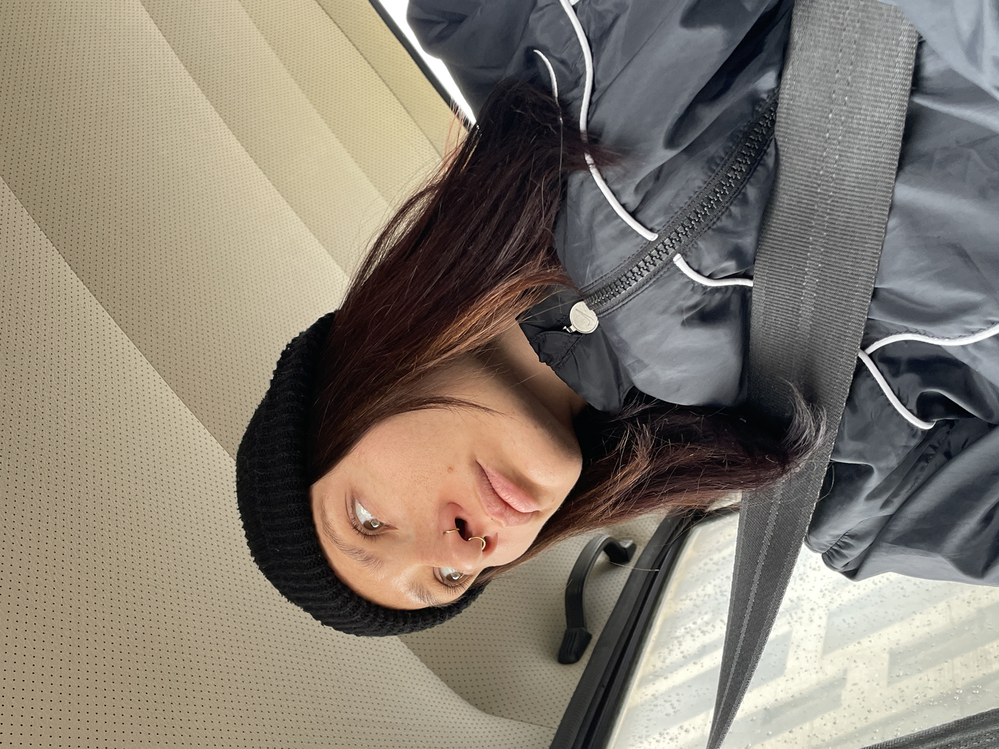 A woman with brown hair and a black beanie is in a car, wearing a black jacket with white piping and a seatbelt. She has a nose ring. The car interior has beige perforated leather seats and a black door handle. Raindrops are visible on the window. There is no information to suggest the location is in Izhevsk, Russia.